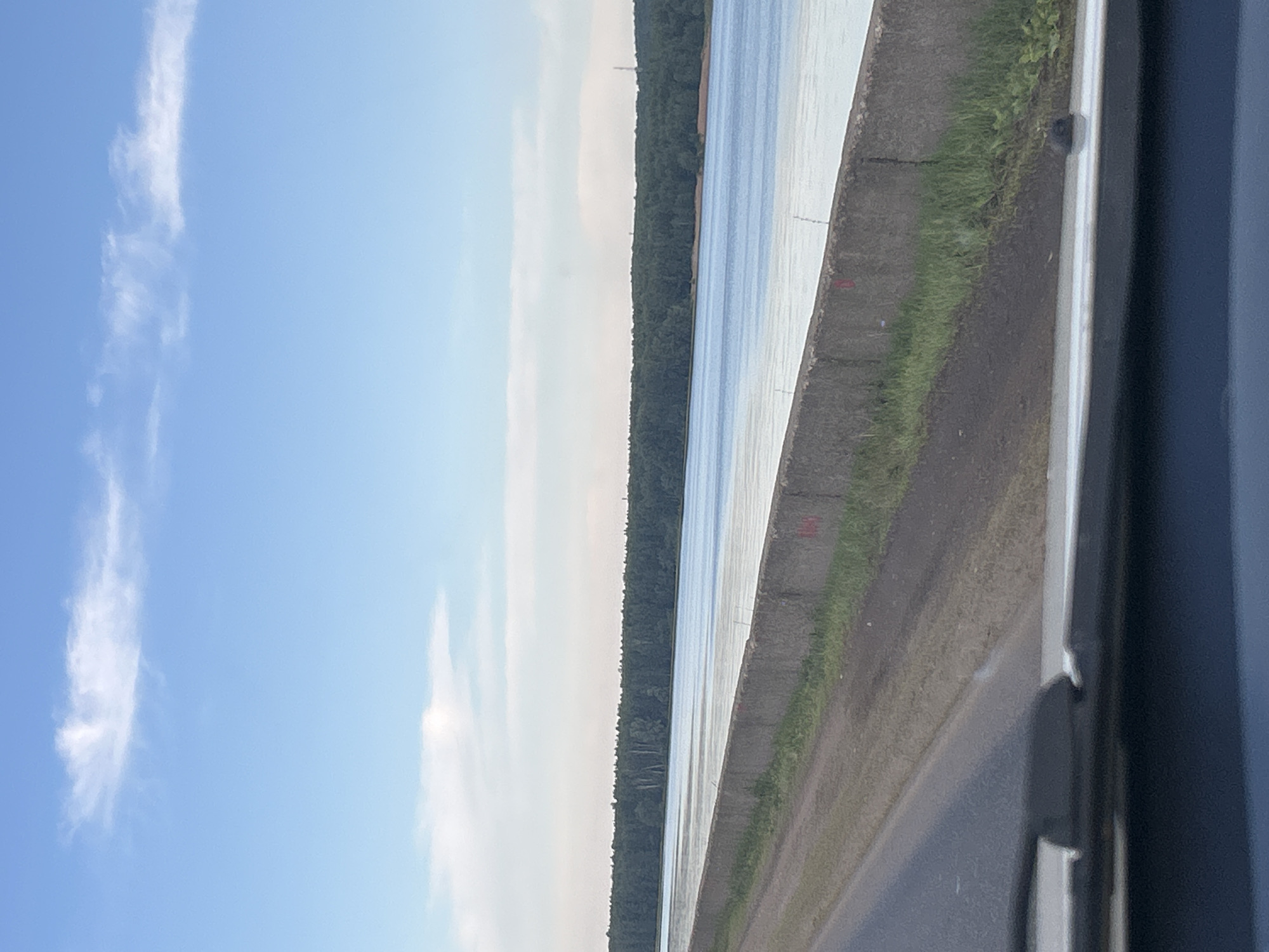 The image depicts a view from a vehicle traveling along a road next to a body of water. The road is paved with asphalt and has a dirt shoulder. A concrete barrier runs alongside the road, separating it from a wide river or canal. Beyond the water, a dense forest lines the opposite bank, with a small section of sandy beach visible. The sky is clear blue with scattered white clouds. The scene appears to be a scenic route, possibly a road leading to or from the city of Chaikovsky, Russia, which is known for its proximity to the Kama River and surrounding forests. The image captures a serene natural landscape, likely during daylight hours.