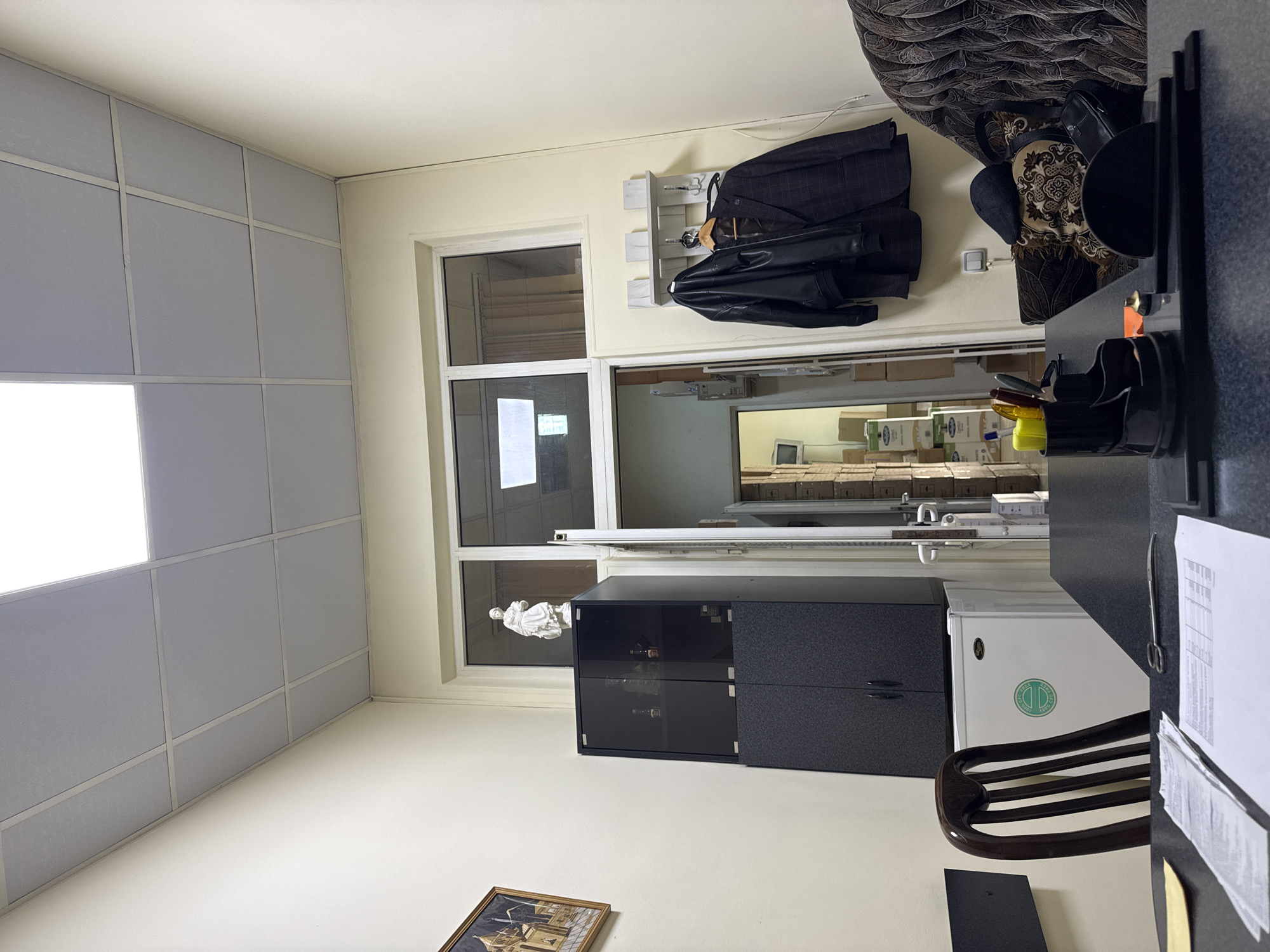 The image shows an interior space, likely an office, with a desk occupying the right side of the frame. On the wall to the right of a doorway are hanging clothes - a suit jacket and a leather jacket on a coat rack.
The doorway leads to an adjacent room filled with stacked cardboard boxes. In the background of the doorway a CRT monitor can be seen.
Below the doorway there is a dark-colored cabinet and a mini-refrigerator. A statue is placed on top of the cabinet.
A picture hangs low on the wall below the cabinet.
The room appears to be brightly lit, with a grid of ceiling panels indicating artificial lighting.
The location is Yerevan, Armenia. No people are visible in the scene.