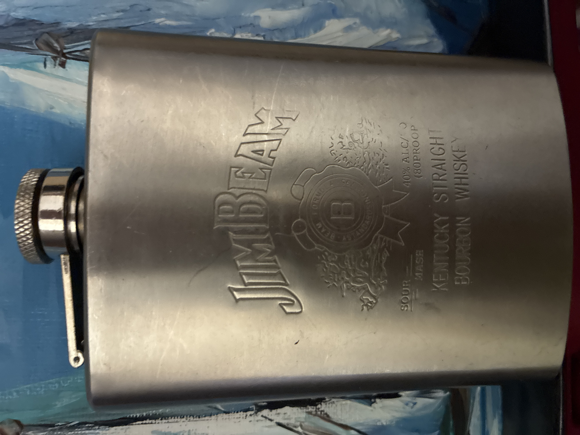 The media file features a metallic hip flask, likely made of brushed stainless steel, depicted horizontally. The flask is prominently branded with "JIM BEAM" in large, embossed capital letters across its surface. The cap of the flask is visible on the left, connected to the body by a small hinged arm.
Detailed embossed text on the flask provides information about the product: "40% ALC/V (80 PROOF)" indicates the alcohol content, and "KENTUCKY STRAIGHT BOURBON WHISKEY" specifies the type of spirit. The words "SOUR MASH" are also visible vertically near the bottom right. A circular emblem is embossed below the "JIM BEAM" logo, featuring the letter "B" at its center and encircled by finer, less legible text, including phrases like "FORMULA" and "BEAM 1795," suggesting historical references to the brand. The flask shows some minor scratches and wear on its surface.
The background consists of a blue and white patterned surface, which appears to be a painting or a textured fabric, possibly depicting abstract waves or clouds. The lighting highlights the metallic sheen and embossed details of the flask. The image focuses solely on the object, with no human subjects or specific activities taking place. This media file is located in Varna, Bulgaria.