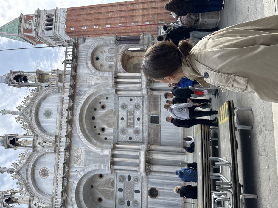 The image depicts St. Mark's Square in Venice, Italy, showcasing the facade of St. Mark's Basilica and a portion of St. Mark's Campanile. In the right foreground, a female individual is viewed from behind, wearing a beige trench coat and with her hair tied in a low bun. She is looking upwards and to the left, observing the architecture.
In the midground, a group of approximately ten individuals are present. Several are seated on elevated wooden platforms with visible metal support structures. One person wears a blue jacket, another a black jacket, both seated. A cluster of young men stand, some appearing to be in conversation.
The background is dominated by the ornate, multi-layered facade of St. Mark's Basilica, characterized by grey and white marble, numerous arches, columns, intricate sculptures, circular medallions, and pinnacles. A large clock face is visible above one of the arches. To the right of the basilica, St. Mark's Campanile rises, a tall red-brick bell tower featuring arched openings near its apex and a distinct green pyramidal roof surmounted by a golden angel statue. A flag displaying the Lion of St. Mark is affixed to the campanile. The ground is paved, and the sky is partly cloudy.