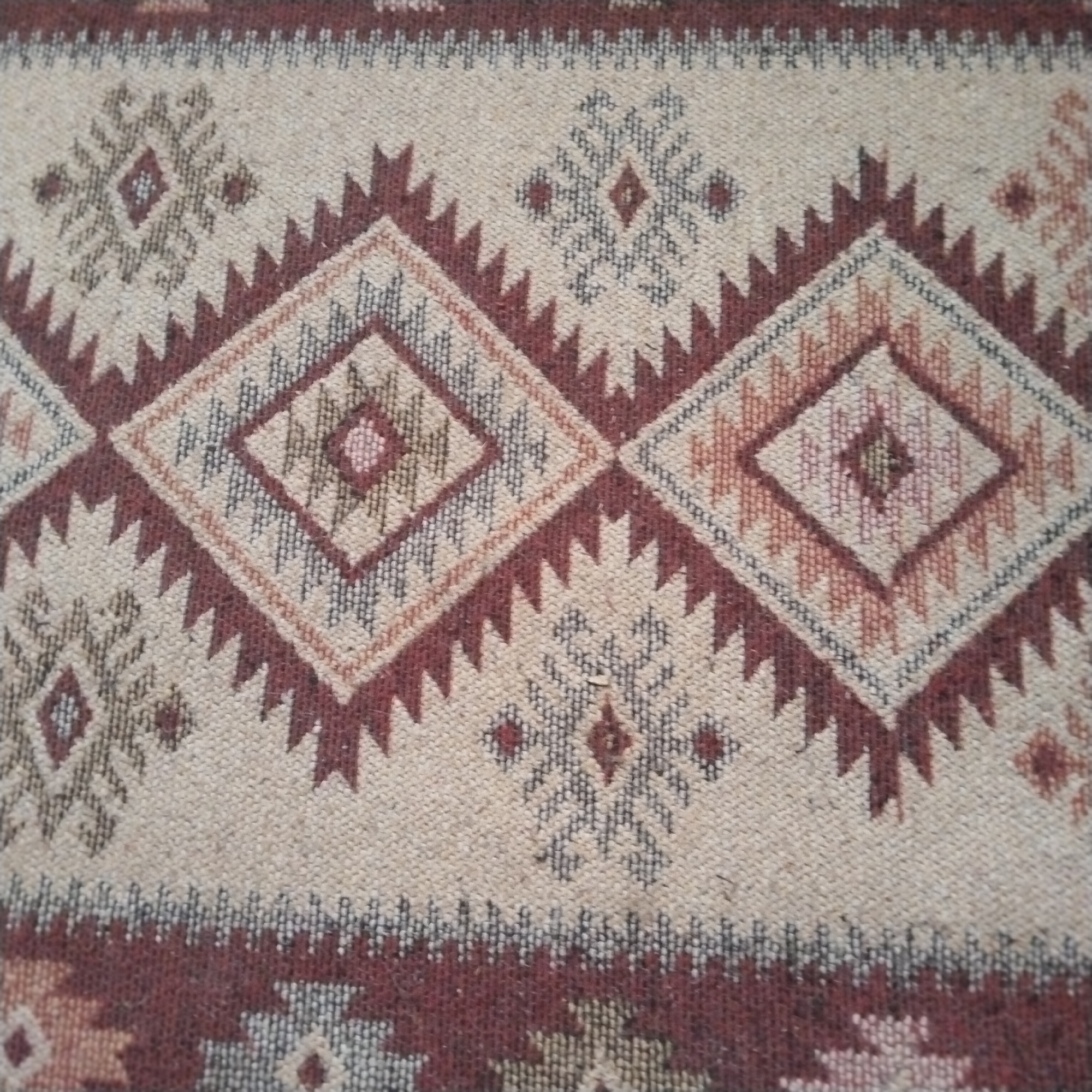 This is a close-up, overhead shot of a woven textile, likely a rug or blanket, displaying a geometric pattern. The primary colors are a neutral beige or cream as the background, accented with shades of maroon, brown, grey, and a hint of pink. The pattern features repeating diamond shapes and zig-zag or sawtooth motifs, characteristic of traditional indigenous weaving styles. The texture of the weave is clearly visible, suggesting a natural fiber material. There are no people, specific objects, or a discernible scene depicted; the focus is solely on the decorative pattern of the fabric. The lighting is even and appears to be artificial, with no indication of time of day or weather conditions. The location context of Kalachinsk, Russia, does not provide any direct visual cues within the image itself. No text is visible.