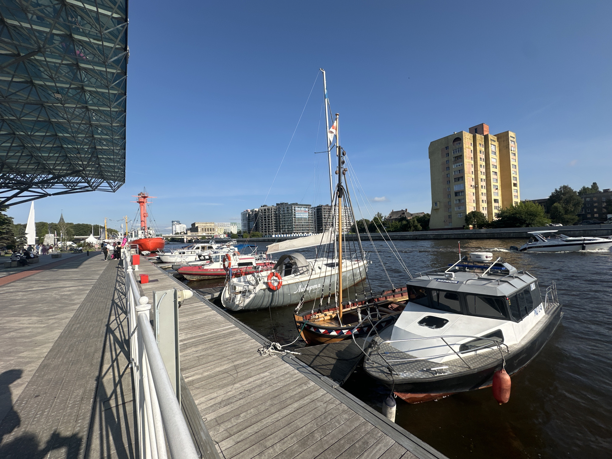 The image depicts a waterfront scene in Kaliningrad, Russia, likely at the Pregolya River embankment. On the left, a modern building with a glass facade dominates the foreground, casting long shadows across a wooden promenade. Several boats are moored along the wooden pier, including a white patrol boat with "ПОЛИЦИЯ" (Police) written on its side, a sailboat with a mast and sails, and a red rescue boat. In the background, a yellow residential building with multiple stories stands out, along with other urban structures and trees. The sky is clear and blue, indicating a sunny day. People are visible walking on the promenade and sitting on benches. The overall impression is one of a well-maintained urban waterfront area.