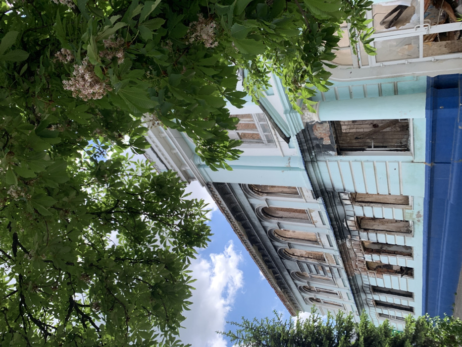 The image shows a building in Izium, Ukraine, located at the coordinates 49.192, 37.279. The building is light blue with white trim and a dark blue base. There are large windows on the side of the building, many of which are broken or boarded up. The building appears to have been damaged by fire, with soot marks on the walls. The image is taken from a low angle, looking up at the building. There are branches of a leafy tree in the foreground. The tree is in bloom and has white flowers. The sky is a light blue with some white clouds. There are no people visible in the image.