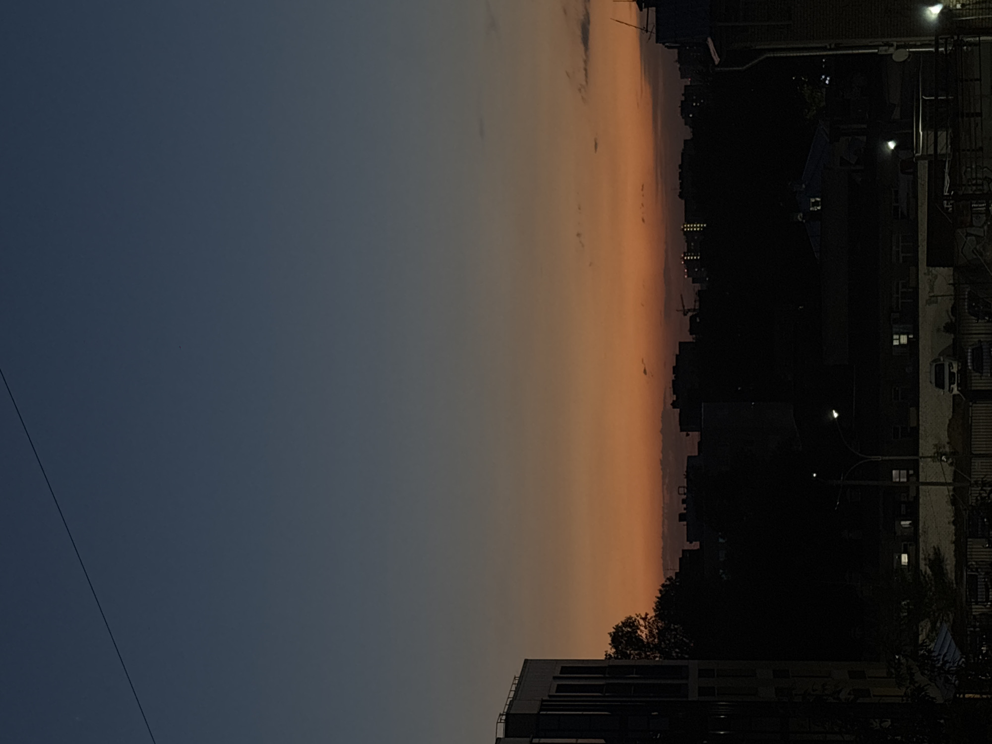 The image depicts an urban landscape at dusk, potentially in Perm, Russia, based on contextual clues. The sky transitions from a deep blue on the left to a warm orange on the right, indicating a sunset or sunrise. Silhouetted buildings and trees form a dark skyline against the colorful sky. Streetlights are visible, illuminating the foreground structures. The image includes utility lines and features of buildings like windows and balconies.