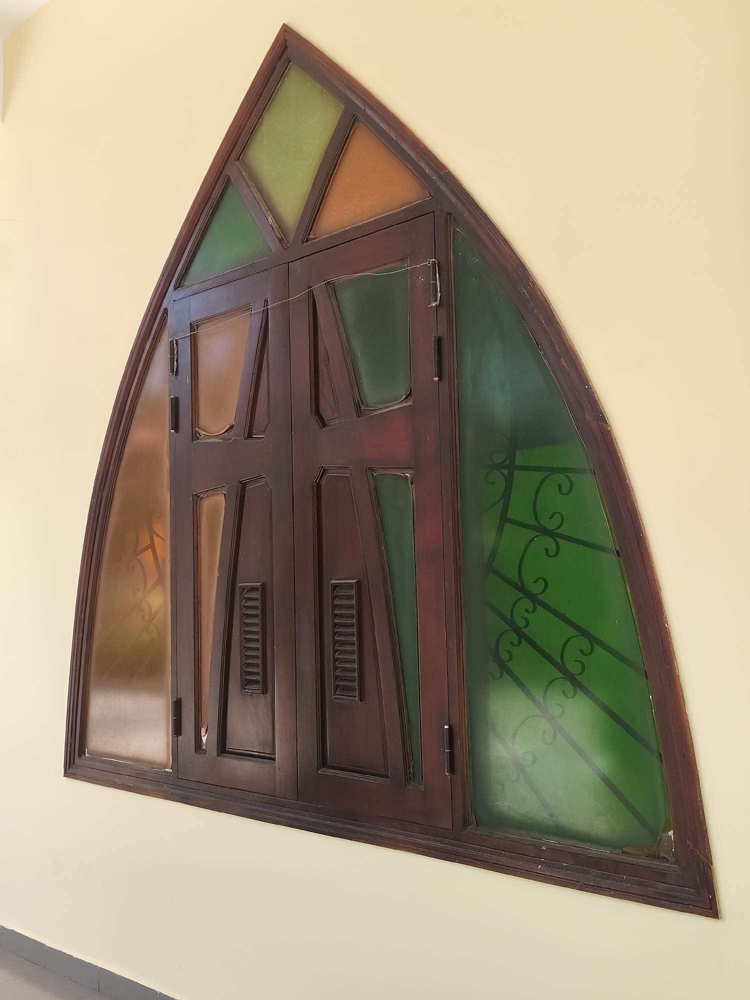 The image shows a close-up, eye-level view of a triangular stained-glass window mounted on a light yellow wall. The window is framed by dark wood. The stained glass is divided into several geometric sections, with colors including green, amber, and a lighter brown. The main body of the window consists of two wooden doors with louvered vents and decorative panels. The doors have hinges visible on the left side. The left side of the window features a section of amber-colored stained glass, while the right side has a large section of green stained glass with decorative black metal grilles. The window appears to be located in a building in Da Lat, Vietnam, although specific architectural context is not provided. The lighting is even, suggesting it is daytime.