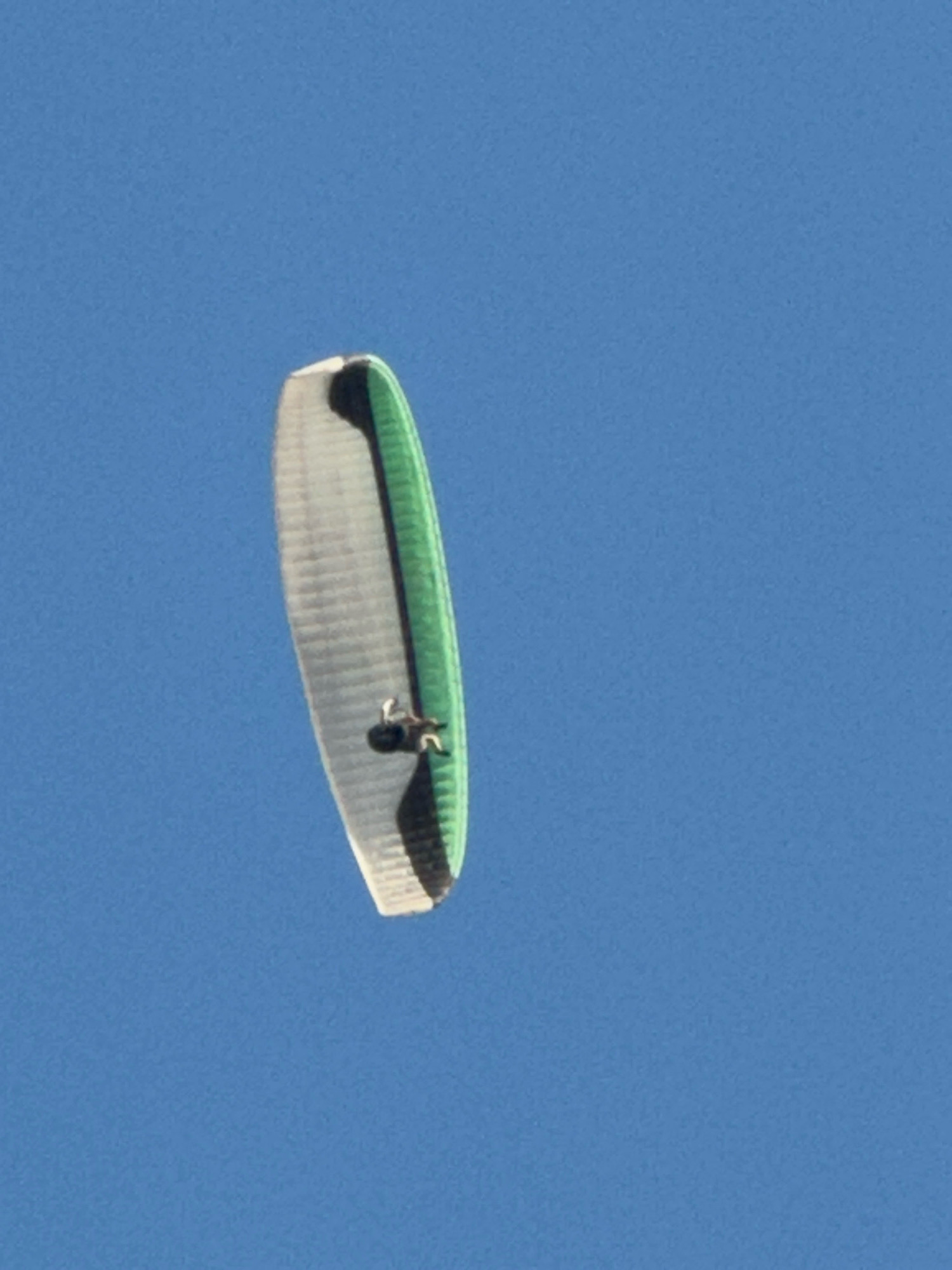 A paraglider with a white and green wing is suspended in a clear blue sky. The paraglider appears to be in flight. There is a person visible on the paraglider, wearing a dark helmet and clothing. The paraglider is positioned vertically in the frame. The background is a solid expanse of blue sky, with no discernible landmarks or geographical features visible, therefore the location in Boreti, Montenegro, cannot be determined from the image alone.
