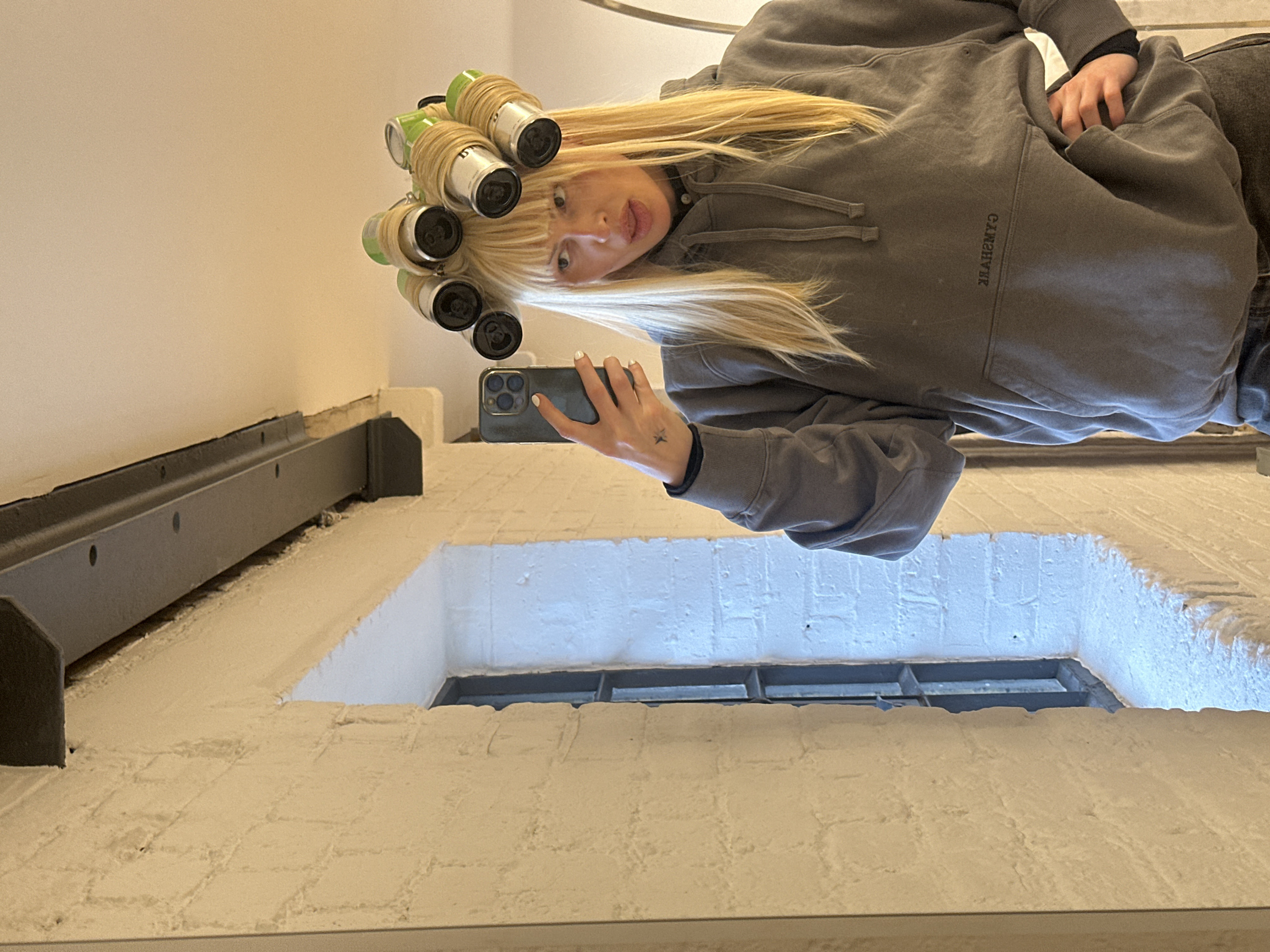 The image is a mirror selfie featuring a person with long, straight blonde hair and bangs, looking directly into the camera with a slightly open mouth, possibly indicating a playful or surprised expression. The person has several hair rollers in the front and top sections of their hair, particularly in the bangs. These rollers appear to be improvised, made from small aluminum cans (some green, some silver) wrapped with a light-colored material, likely string or tape, to hold the hair in place.
The individual is wearing a grey hooded sweatshirt, with a dark long-sleeved garment visible underneath the sleeves. On the back of the left hand, a small, dark star-shaped tattoo is visible. The person's fingernails are painted white. They are holding a dark-colored smartphone, which has a distinct multi-lens camera system, in their left hand to capture the selfie.
The background, which is the reflection of the room behind the person, shows an indoor environment. A plain, light-colored wall (possibly off-white or beige) is visible. Below this wall, there is a dark, metallic-looking structure that runs horizontally, featuring several small circular holes. Further down, there is a lower wall section with a distinct textured appearance, suggesting painted white bricks. Within this white brick section, a dark, rectangular opening, likely a window, is visible, containing a grid-like internal structure. Text is present on the front of the grey hooded sweatshirt, which is mirrored and appears to read "YRBMAƆ" or "CAMBRAY" in reverse. The overall lighting suggests an indoor setting.