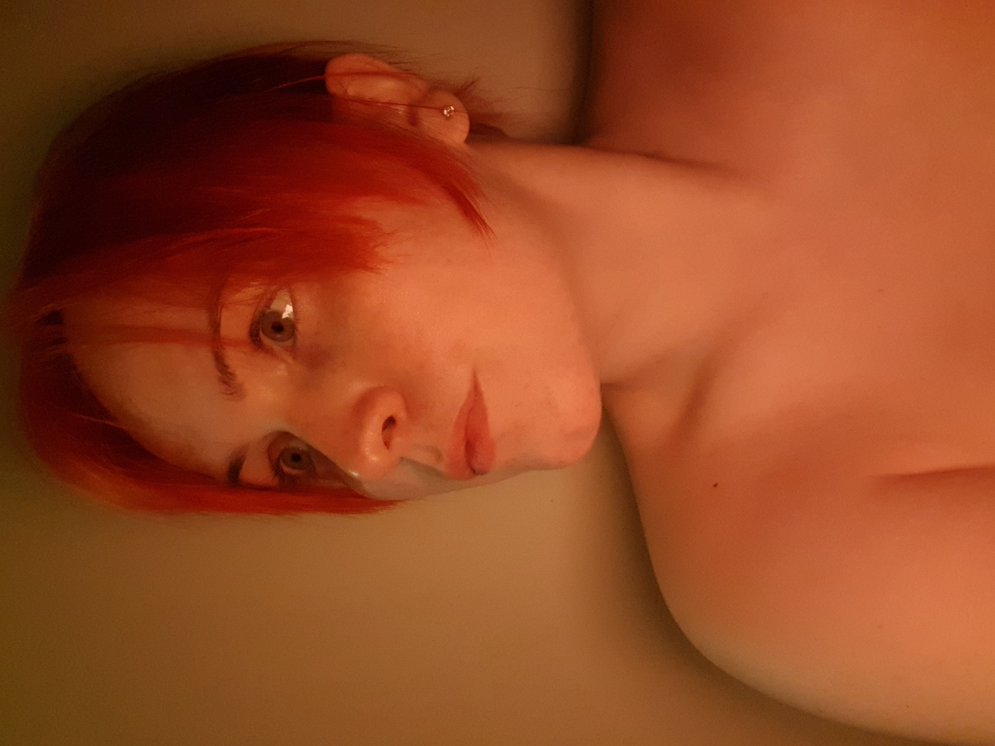 The image presents a close-up of a person with red hair partially submerged in water, possibly a bathtub, filled with a yellowish-brown liquid. The person's head is tilted to the left, and their gaze is directed downward and to the left. A small earring is visible in the left ear. Skin tones are warm due to the lighting, which is orange and somewhat subdued. The environment suggests an indoor setting. The location in the city is unknown, unknown.