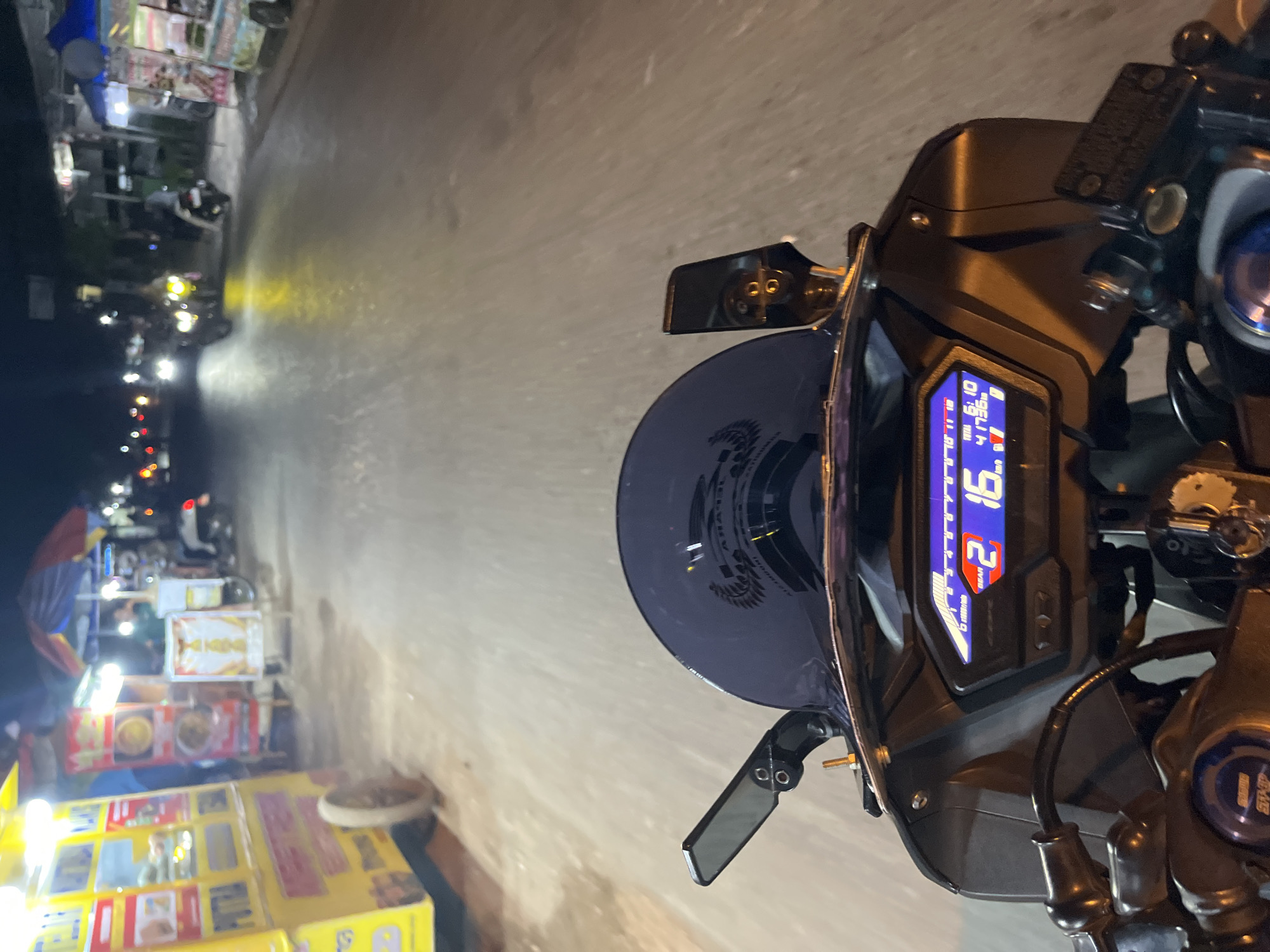 The image captures a first-person perspective from a motorcycle at night, navigating a street in Pecangaan, Indonesia. The road is dimly lit, with streetlights and the headlights of other vehicles casting a yellow glow. On the left side of the road, there are several food stalls, one prominently featuring a yellow cart with the word "PUTRA" visible. These stalls are illuminated by bright lights, illuminating their wares. In the distance, other motorcycles are visible, some with their headlights on. The motorcycle's digital dashboard displays the gear as "2" and the speed as "16". The environment appears to be a busy street with local businesses operating into the night.