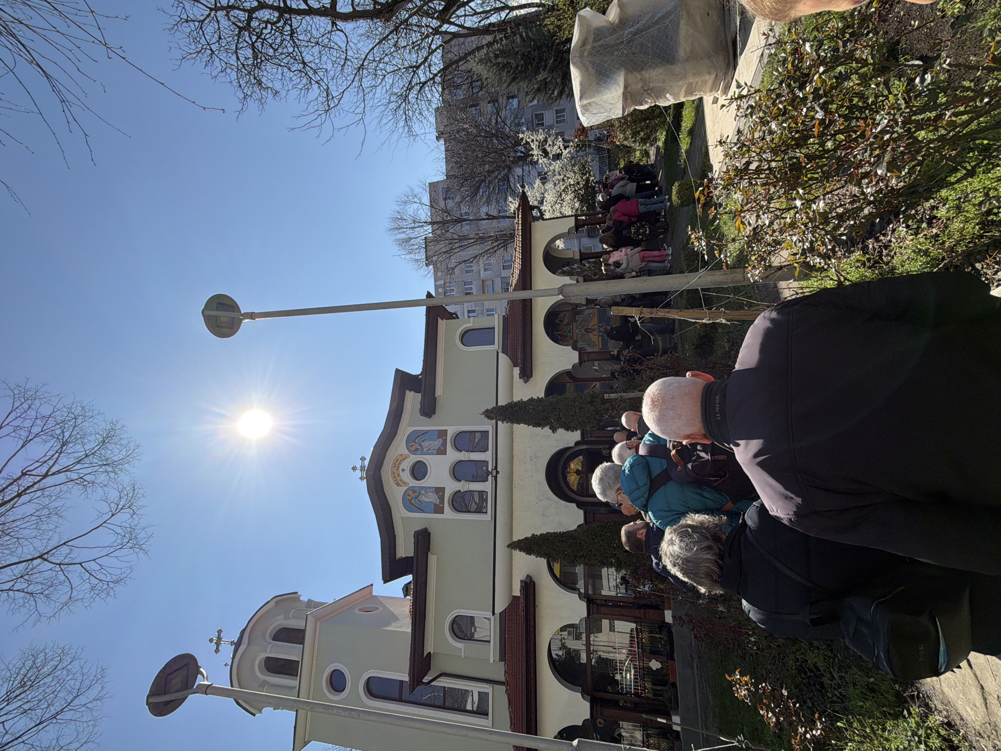 The image shows a low-angle, daytime view of a group of people congregated outside the Saint George Rotunda Church in Sofia, Bulgaria. The church, with its light-colored facade, visible religious iconography, arched doorways, and windows, is the primary structure in the image. Some bare trees are visible. A light pole stands in front of the church with a round reflector. In the background, there is a glimpse of a tall, residential building. The sun is shining brightly. The people are standing and some are facing the church. Some of the people are positioned in the foreground, with their backs towards the camera. There is also some foliage and shrubbery near the foreground.