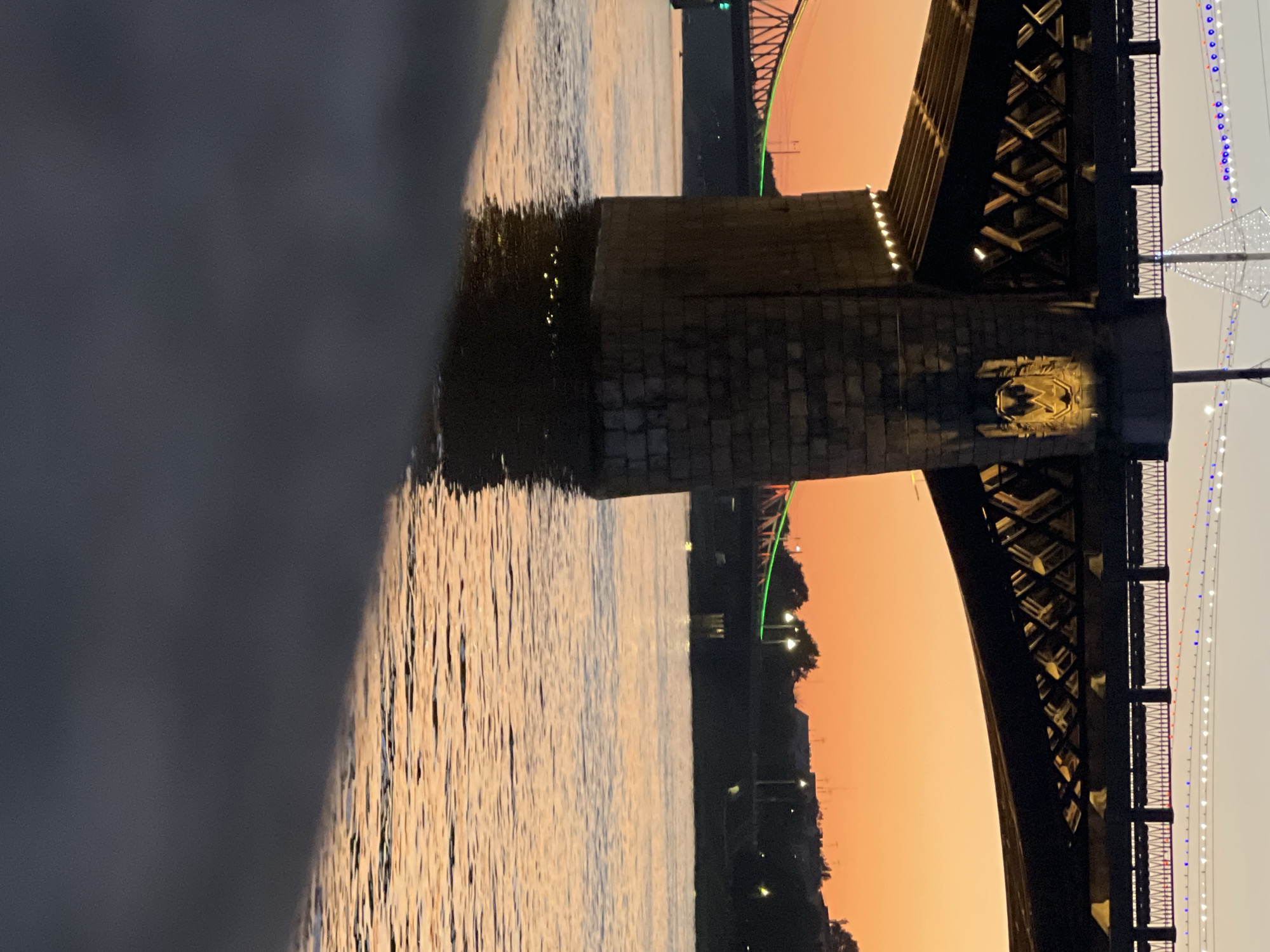 An eye-level, medium shot captures a river beneath a bridge at sunset in Tver, Russia. The sky is a gradient of orange and yellow, with a faint white structure illuminated by decorative lights against the horizon. A large, stone bridge pier dominates the center foreground, its rough-hewn blocks darkened by water that laps at its base, reflecting the warm hues of the sky. The underside of the bridge features a series of diagonal beams and is illuminated by small, warm lights. To the left, another bridge is visible, adorned with a green light. Silhouetted trees and buildings line the far bank of the river. The water itself is choppy, rippled, and reflects the colors of the sunset. A dark, blurry foreground element suggests a walkway or railing in the immediate foreground. No people are visible in the scene.