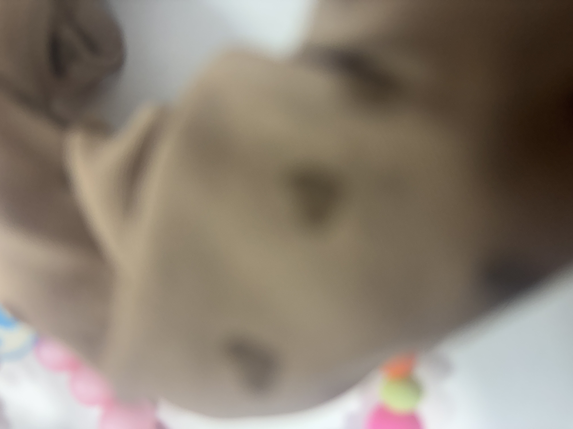 The image displays a severely out-of-focus scene centered on a light brown or tan, smooth surface. Multiple darker, indistinct heart-shaped patterns or markings are visible on this surface, with at least three clearly discernible. The background and left periphery contain highly blurred elements, including indistinct spherical or rounded shapes in shades of pink, green, orange, and light blue, against a bright, undefined background. No identifiable figures, specific actions, or environmental details are discernible. The location is undeterminable.