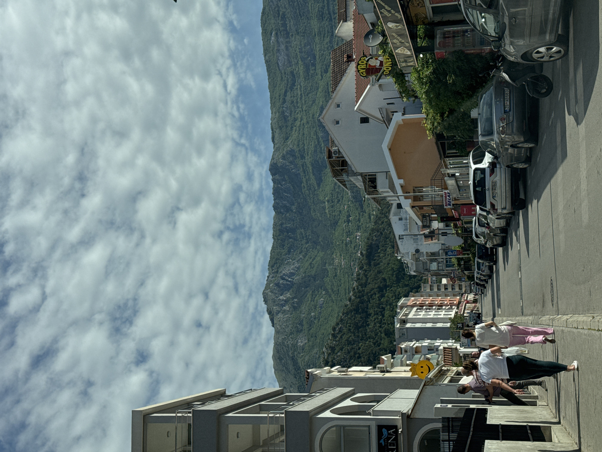 A daytime, street-level shot shows a narrow, one-way street in the city of Budva, Montenegro. The street is lined with parked cars on the right and modern buildings on the left. Three people are walking along the sidewalk: a woman in a white shirt and dark pants, another woman in a white shirt and pink pants, and a man whose face is not clearly visible, wearing a purple shirt. Businesses with signs in English and other languages can be seen along the street, including one for "GYROS DONER PIZZA." In the background, a steep, green mountain rises up behind the buildings. The sky is light blue with scattered clouds. The image is rotated counterclockwise.