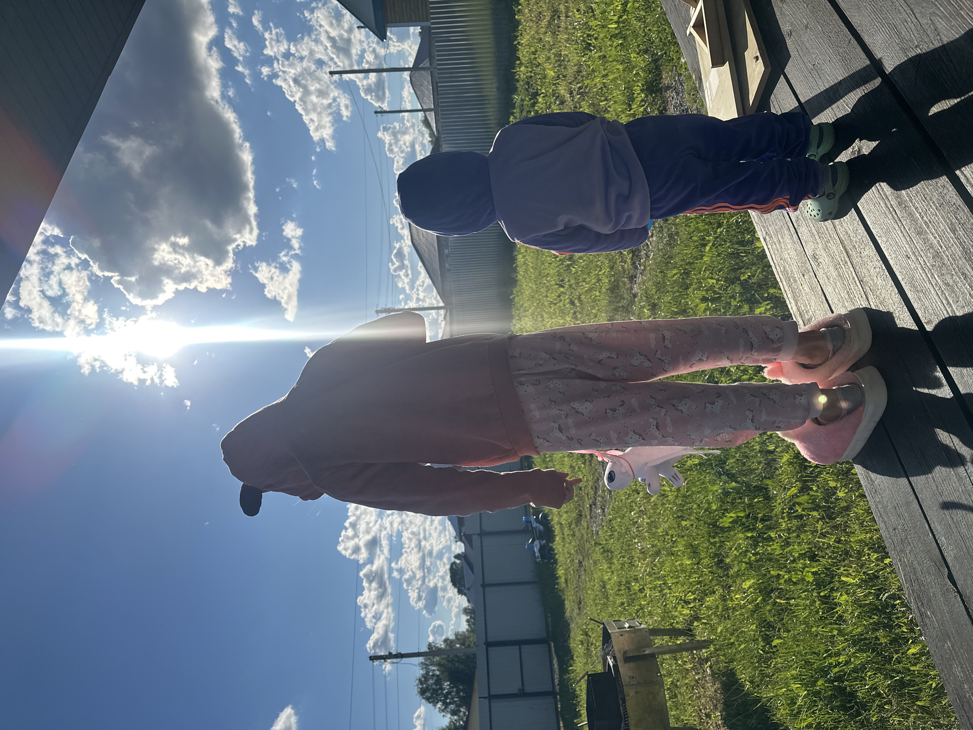 Two children stand on a dark, wooden deck. One child is wearing a red hooded sweatshirt and pink pajama pants with a unicorn pattern. They have pink fuzzy slippers and hold a small, plush unicorn. The second child wears a purple hooded sweatshirt and purple sweatpants with an orange stripe. They are wearing green Crocs. The backdrop features green grass, a metal fence, and a blue sky with scattered white clouds. A bright sun is visible, causing lens flare. The location is Kharnavy, Russia.