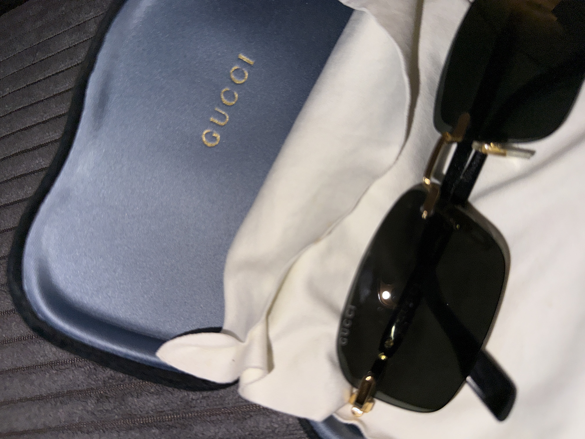 The image shows a pair of black Gucci sunglasses placed on a white cloth, which is inside a blue Gucci case. The case has the Gucci logo embossed in gold. The sunglasses have gold accents on the arms and temples. The background is a dark, textured surface, possibly a piece of furniture. The scene is dimly lit, suggesting it might be indoors. There are no people visible in the image. The location of this scene is not determinable from the image.