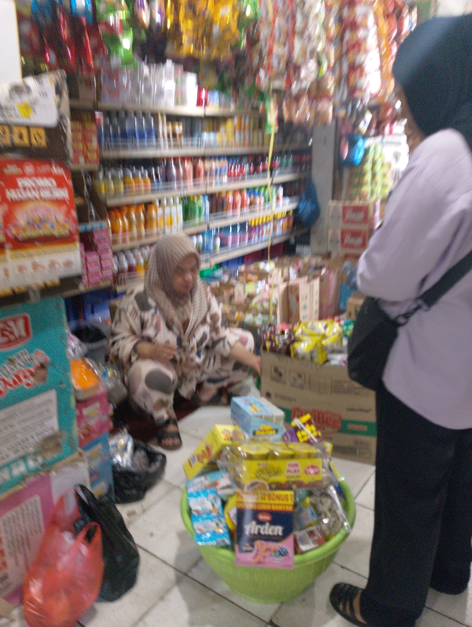 The image depicts an indoor scene within a small shop, likely a convenience store or "warung," located in Lembang, Indonesia. The shop is packed with various goods displayed on shelves and in baskets, suggesting a commercial environment. A woman wearing a hijab and patterned clothing is crouched on the floor, appearing to be organizing or interacting with items in front of her. To the right, another person, also wearing a hijab and a light-colored top and dark pants, stands with their back to the camera, carrying a dark bag over their shoulder. The overall impression is one of a busy, cluttered retail space, with merchandise filling the aisles and shelves.