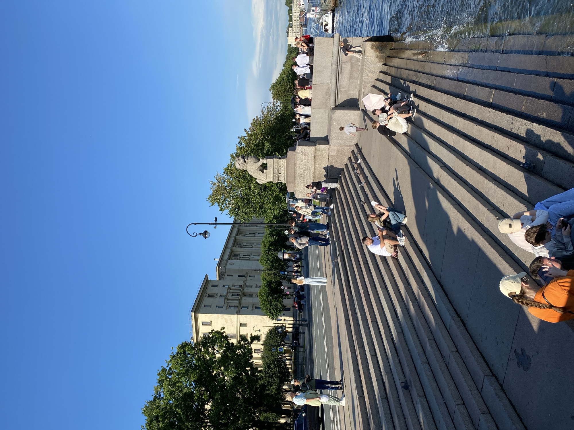 The image depicts a sunny day at the St. Petersburg embankment. A wide set of granite stairs descends to the Neva River. People are scattered across the stairs, some sitting and talking, others looking at their phones, and a few are standing. In the background, a large building with multiple floors and a distinct architectural style stands under a clear blue sky. Trees line the embankment, and a street with cars and pedestrians is visible beyond the stairs. A prominent stone lion statue on a pedestal is located on the upper level of the embankment, overlooking the river. Boats are moored along the riverbank, and the water is calm with some ripples. The scene conveys a relaxed atmosphere, with individuals enjoying the riverside.