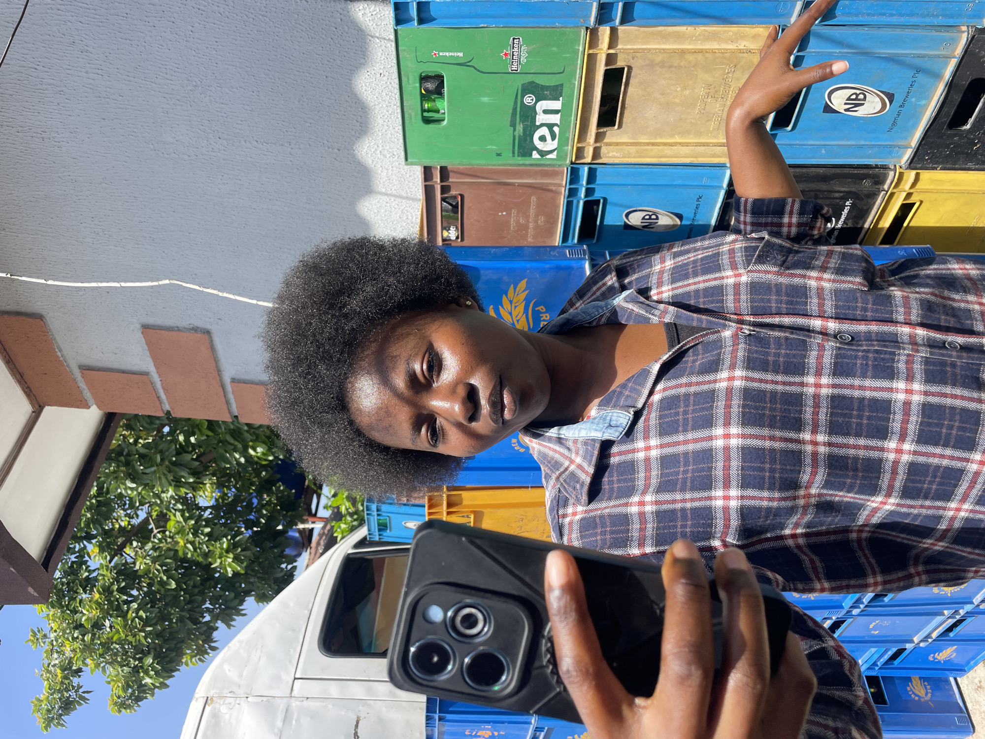 A young Black woman with natural afro hair, wearing a dark blue plaid shirt with red and white accents, is taking a selfie with her smartphone. She is standing outdoors, with a white van visible behind her and to the left. The background consists of stacks of colorful plastic crates, predominantly blue, yellow, and green. Some of the crates have visible branding, including "ken" and "Heineken." The environment appears to be an urban setting, likely in Jos, Nigeria, given the context provided. The lighting suggests it is daytime with a bright sun, casting shadows on the wall behind the woman. The woman is looking at her phone screen, with her left hand holding the device and her right arm extended towards the crates.
