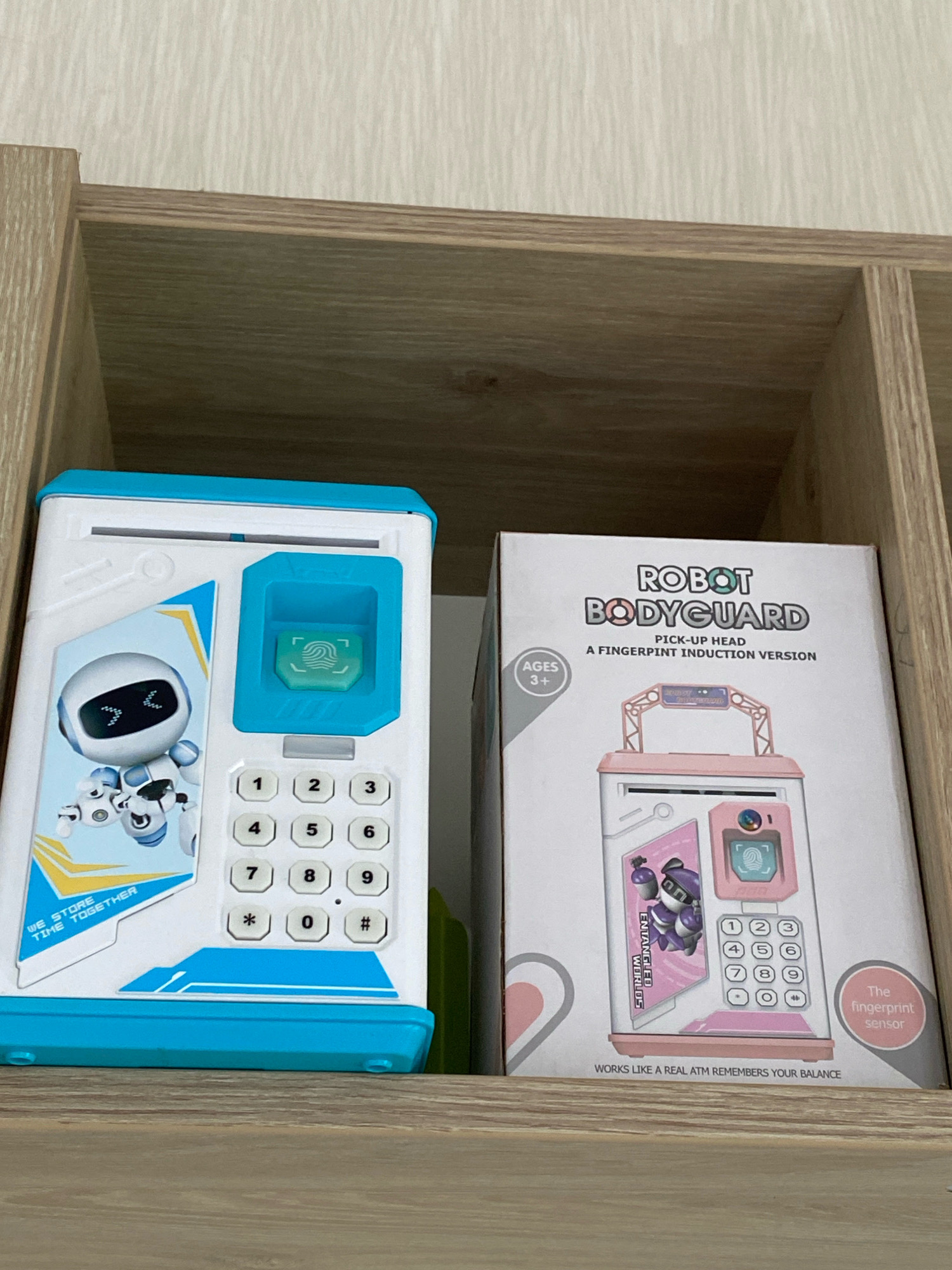 The image shows two items on a wooden shelf against a textured wall. On the left is a white and blue toy ATM with a cartoon robot graphic, a fingerprint scanner, and a keypad. On the right is a pink and white box for a "ROBOT BODYGUARD PICK-UP HEAD A FINGERPRINT INDUCTION VERSION" toy. The box has graphics of a robot and a pink ATM-like device. The environment appears to be indoors, possibly a home or store, with no specific identifiable landmarks of Chișinău, Moldova.