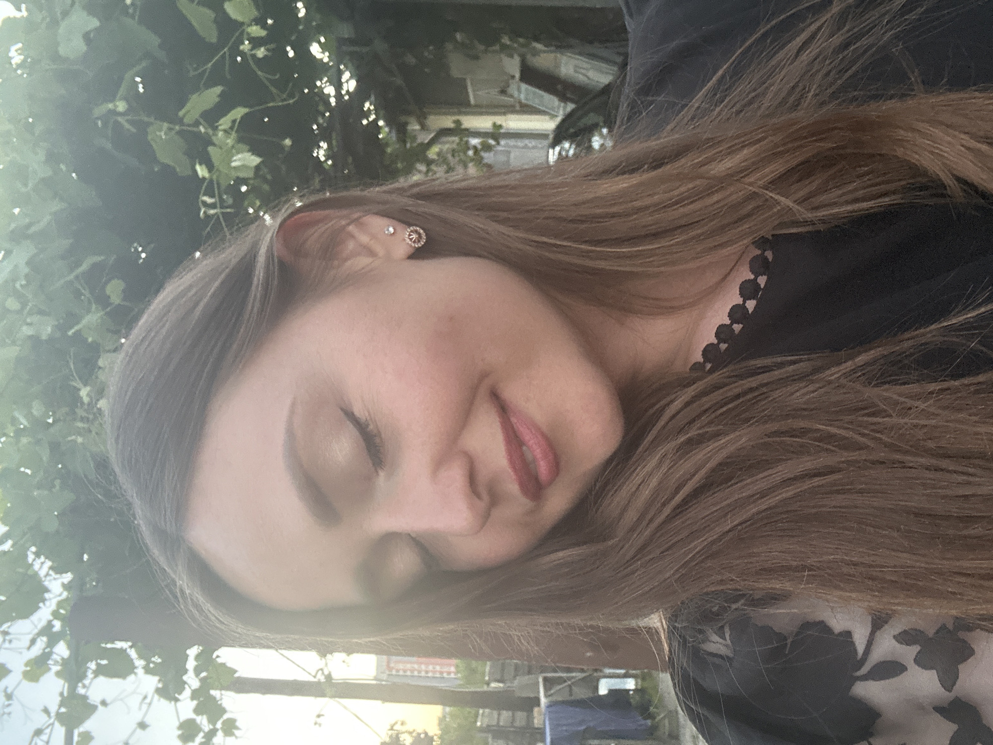 A medium shot shows a young woman with light brown hair and closed eyes, smiling slightly. She is wearing a black top with a scalloped neckline and a floral print on the sleeves. A small, diamond-like stud earring and a rose gold stud earring with a circular design are visible in her right ear. The background suggests an outdoor setting, possibly a yard or garden, with lush green foliage overhead and a glimpse of buildings and a car in the distance. The lighting indicates it is late afternoon or early evening, with the sun casting a soft glow. The location is identified as Bila Tserkva, Ukraine, though specific landmarks are not identifiable from the image.