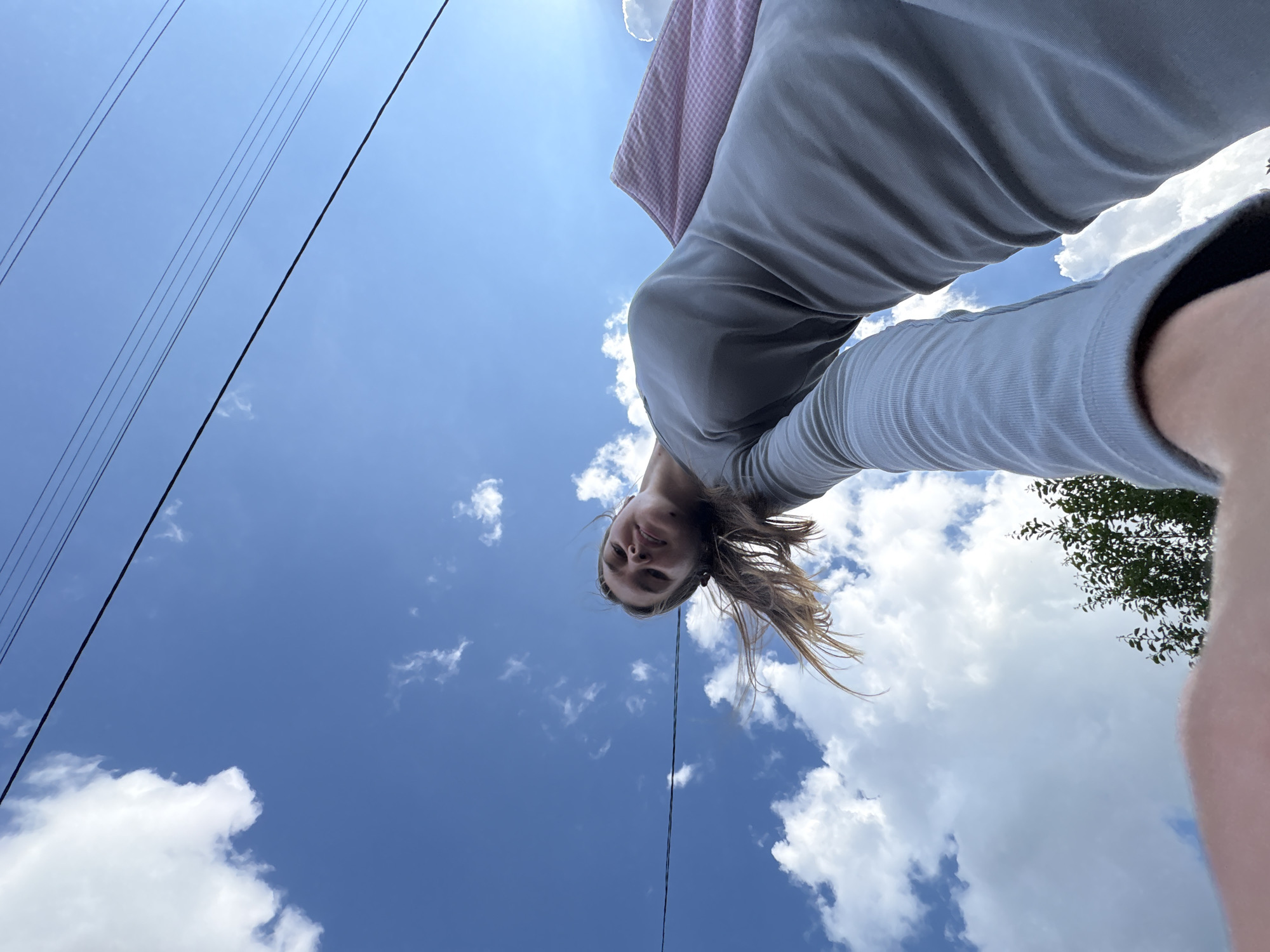 An outdoor, low-angle, wide shot shows a young woman looking up at the camera against a bright blue sky with scattered white clouds. She is wearing a light gray long-sleeved dress and her hair is brown and windswept. Her right arm is extended towards the camera, with her hand out of frame. A pink and white checkered bag is draped over her left shoulder. Electrical wires are visible against the sky in the upper portion of the frame. The lower right corner of the image shows a small patch of green foliage, suggesting the presence of trees. The location is identified as Primorskiy, Russia.