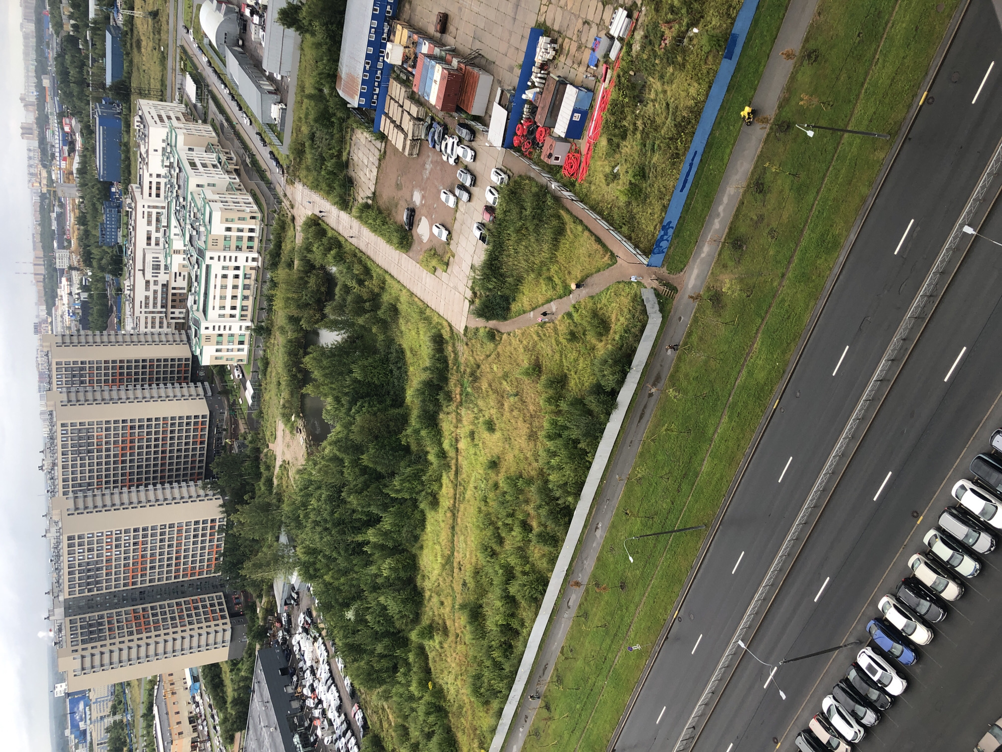 This is an aerial, high-angle, full shot image of an urban landscape under a cloudy sky. The scene is dominated by modern high-rise apartment buildings with beige and grey facades and numerous windows, some with orange accents. In front of these buildings are lower, white and green multi-story structures, possibly offices or commercial buildings, with a patterned facade.
A large green space with dense trees and some areas of dried grass is situated between the buildings and a paved pathway. Several vehicles are parked on a street in the foreground, including a row of sedans and SUVs. Another area with many parked cars is visible near a large, dark grey building.
Further into the scene, a construction or industrial area is present, with stacks of shipping containers, building materials, and pipes. A paved walkway with a light blue fence borders a green grassy area. A person is walking on the paved area near the fence, and a yellow ATV is being ridden on another paved path. The road in the foreground is wet, suggesting recent rain. The overall environment is a mix of residential, commercial, and industrial development within a city.