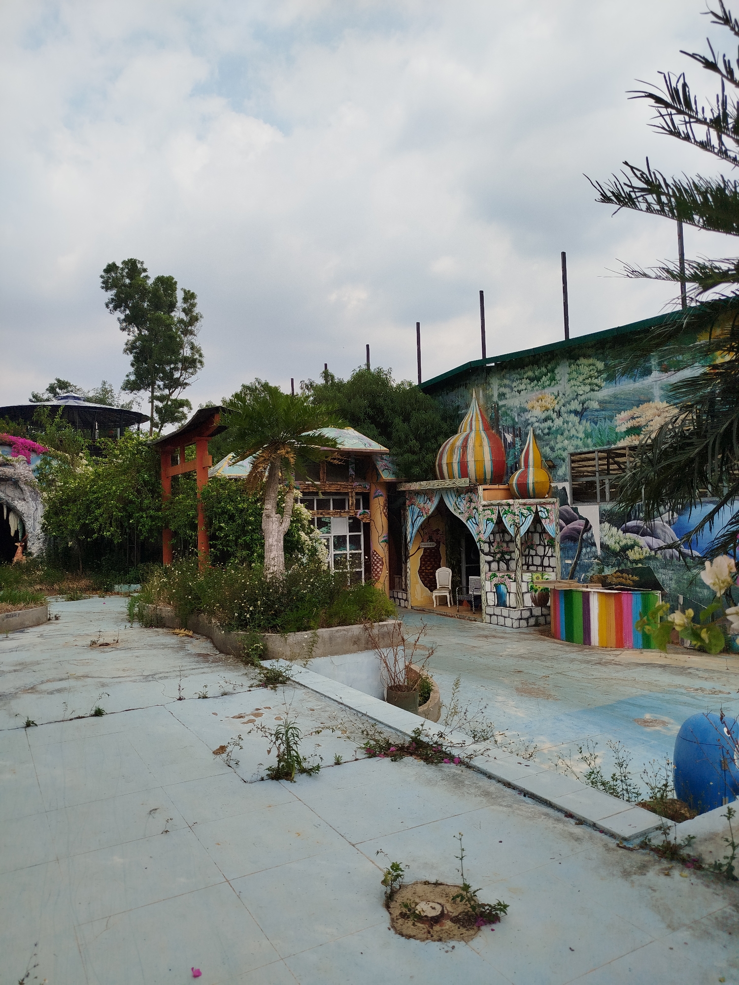 The scene depicts a neglected, possibly abandoned, amusement or tourist attraction in Da Lat, Vietnam. The ground is a light blue, tiled or painted surface with cracks and weeds growing through the gaps. Several small structures are visible, including one with a red arched gate, another resembling a miniature dilapidated house, and a third structure with colorful domes reminiscent of a fairytale or Middle Eastern design.
Behind these structures is a larger building with a faded mural of a landscape. The structures appear overgrown with vegetation. A blue decorative object is visible in the foreground. The sky is overcast. There are no people visible. The overall impression is one of decay and abandonment.