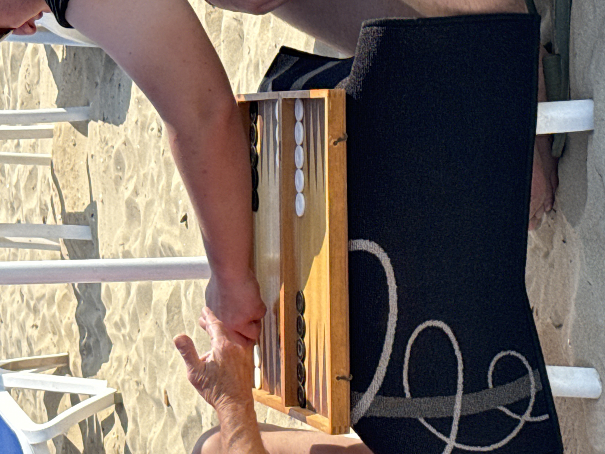 The media file shows a close-up, overhead view of two people playing backgammon on a beach. The game board, made of wood, is set up with black and white checkers. One person's arm and hand are visible, reaching towards the game board. The sand is a light beige color and appears slightly wet, with ripples suggesting it is near the water's edge. White lounge chairs and their supports are visible in the background, along with a large black towel with a decorative white pattern. The lighting indicates it is a sunny day, casting shadows on the sand. The overall scene suggests a relaxed, leisurely activity taking place in an outdoor beach setting, likely during daylight hours. The location is identified as Sozopol, Bulgaria. There is no visible text in the media file.