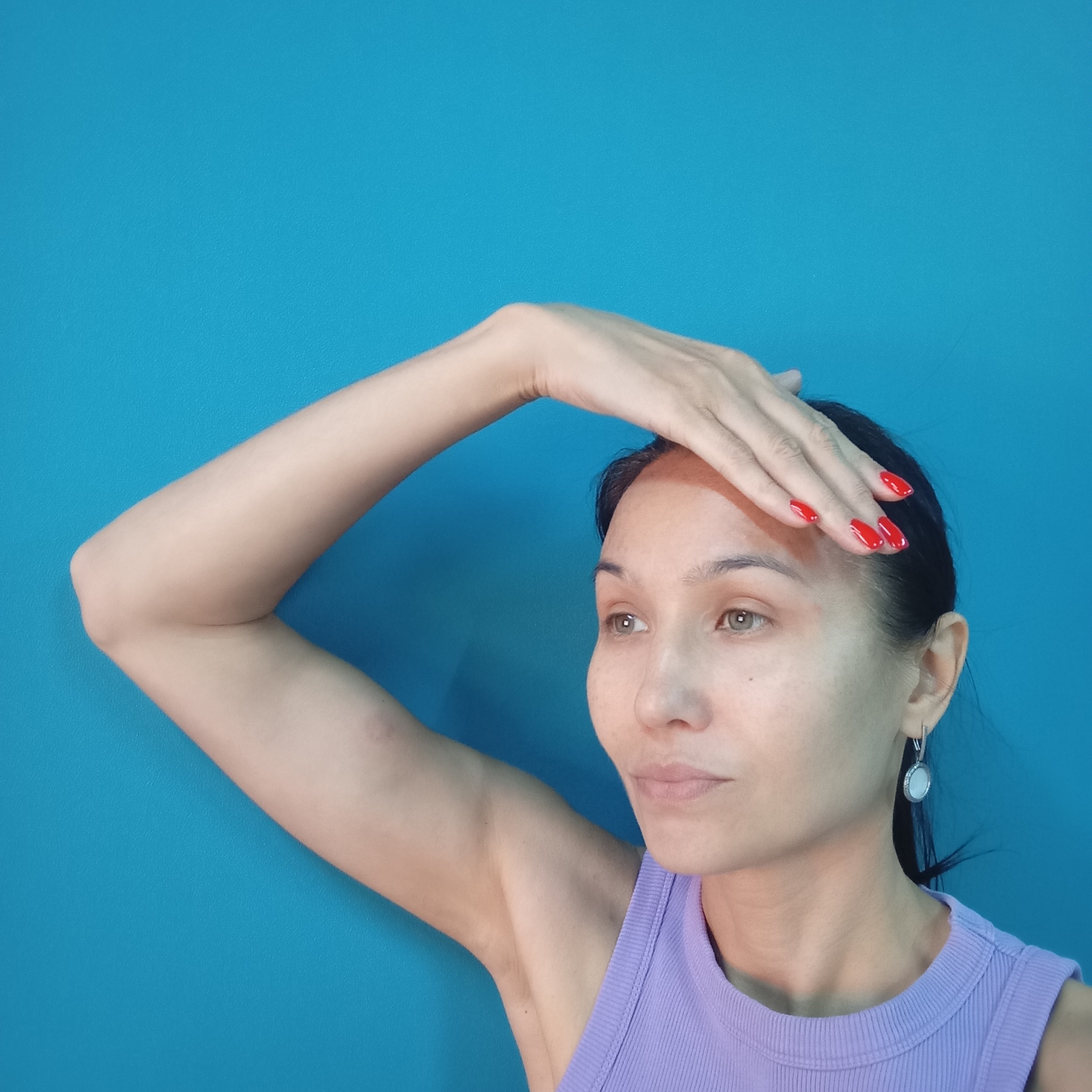 A medium shot shows a woman with dark hair and light skin against a bright blue, textured background. She is looking to her right, with her right arm bent and her hand resting on her forehead. Her nails are painted bright red. She is wearing a purple tank top and a silver earring with a white stone in her left ear. The scene is likely indoors, given the smooth, solid background and artificial lighting. There is no visible indication of the city of Pervomaysky, Russia, in the image.