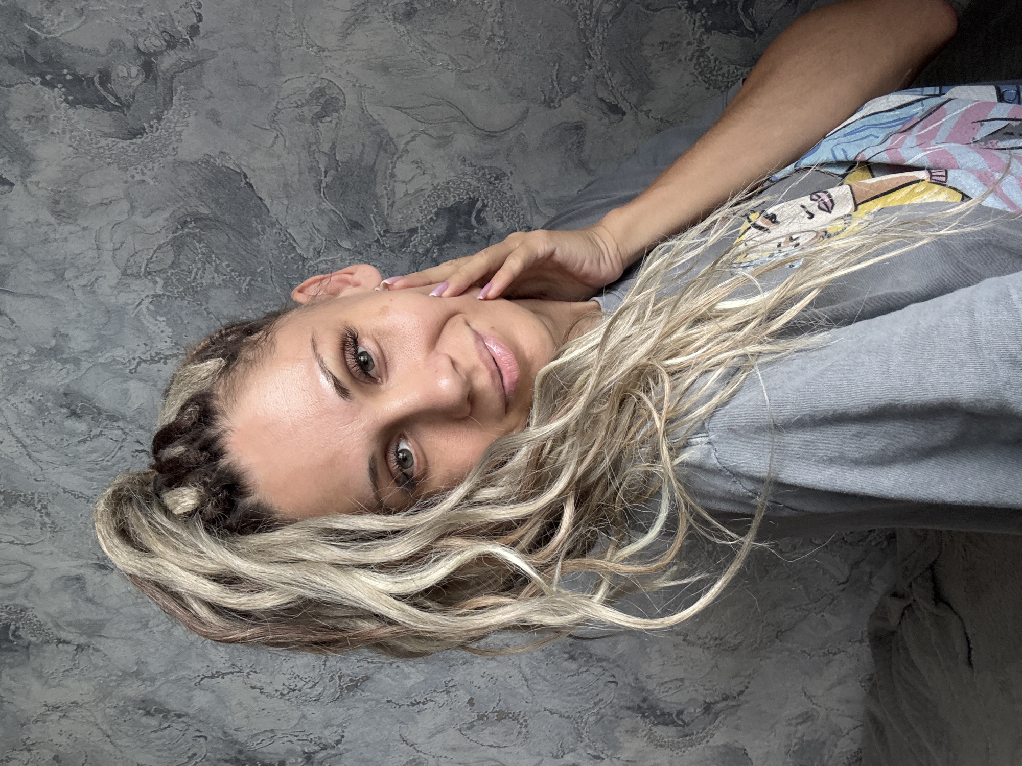 A medium shot of a woman with blonde and brown braided hair styled in a high ponytail. She is wearing a gray t-shirt with a colorful graphic print of a face. Her left hand is resting on her cheek and jawline. The background is a textured gray wall. The image is a close-up, and the woman is looking towards the camera with a slight smile. The woman's skin appears to have some freckles and discoloration. The lighting is soft, and there are no visible shadows. The scene is indoors, and the environment suggests a casual setting. The location is specified as Rusanovo, Russia, although this cannot be confirmed from the visual information alone.