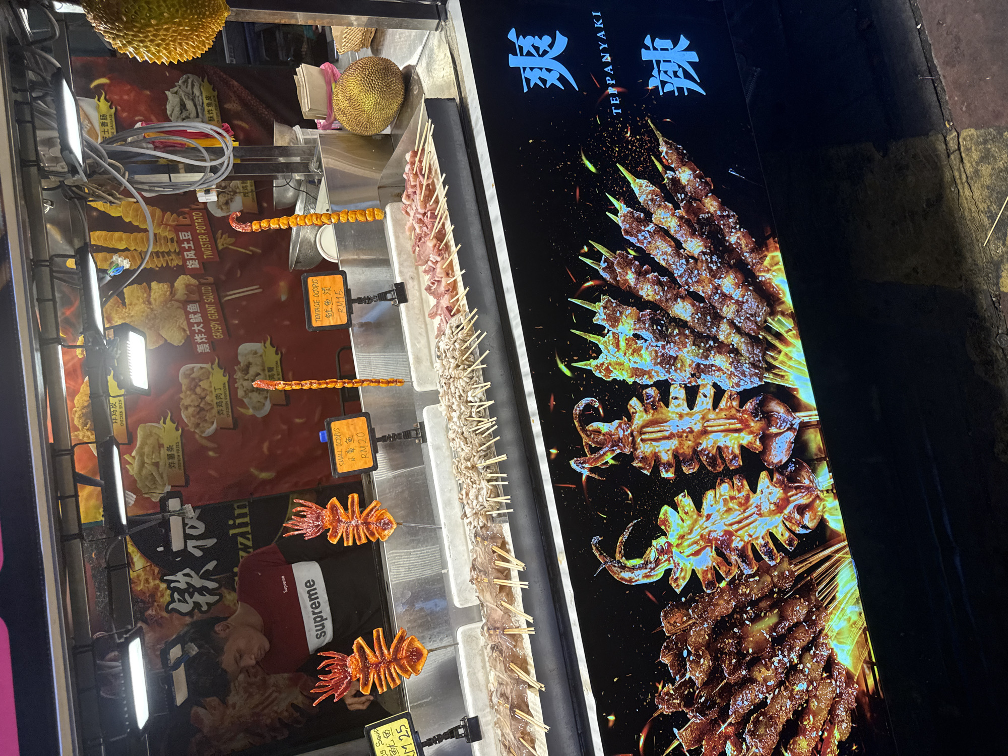 The image captures a vibrant night market food stall, brightly illuminated at night, likely in Kuala Lumpur, Malaysia, as indicated by the "RM" (Malaysian Ringgit) currency. The stall offers a variety of grilled and deep-fried snacks.
On the left side of the stall, several posters advertise different fried items, including "TWISTER POTATO," "CRISPY GIANT SQUID," "FRENCH FRIES," "CHICKEN SKIN," and "CHICKEN POPCORN." A large durian fruit is visible at the top, suggesting it's also available. Prices are displayed for some items, such as "CRISPY GIANT SQUID RM25," "TENTACLE OCTOPUS RM15," and "SMALL OCTOPUS RM20." A person wearing a red "Supreme" hoodie is partially visible behind the counter, near the deep-fried food displays.
In the center, long trays showcase rows of uncooked or marinated skewers, appearing to be small octopus or squid, neatly arranged for grilling. Adjacent to these are skewers of deep-fried spiral potatoes.
The right side of the image features a large, striking LED display sign. This sign advertises "TEPPANYAKI" (铁板烧) in bold Chinese characters and English, showcasing tempting images of various grilled skewers, including meat kebabs and large grilled squid or octopus, with vibrant flames adding to the appeal. Other Chinese characters like "爽" (shuǎng, meaning cool/refreshing) and "辣" (là, meaning spicy) are also visible on the signs. The artificial lighting, combined with the dark background, clearly indicates a nighttime setting.