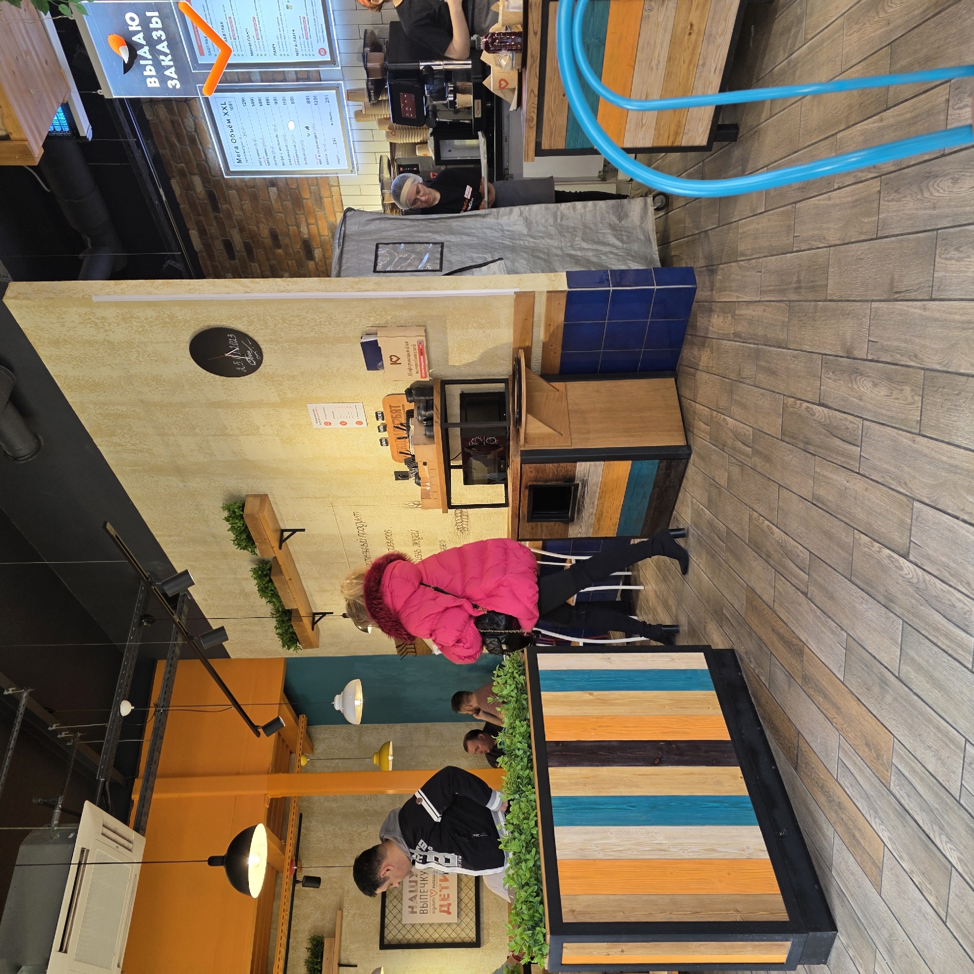 The image captures the interior of a modern and vibrant cafe or fast-food establishment, likely in Saint Petersburg, Russia. The setting features light wooden floor planks and textured cream-colored walls, contrasted with sections of dark blue tiles and bright orange/yellow accents on walls and ceilings.
Several individuals are present. Behind one counter, a staff member in a black top and hairnet appears to be working, possibly preparing orders. Near another counter, another staff member in a black top and apron is visible. A customer wearing a bright pink puffer jacket stands facing a counter, while another man in a black and white jacket stands near a table adorned with artificial greenery. In the background, two other men are seated at a multi-colored striped table.
The cafe's design incorporates varied materials and colors, including tabletops made of horizontally striped wood in shades of brown, orange, and blue. Bright blue flexible tubing or pipes are a distinctive decorative element, running along a wall. Overhead, modern black and white pendant lights illuminate the space.
Visible text in Russian includes a prominent sign that reads "ВЫДАЮ ЗАКАЗЫ" (Issuing Orders), indicating a pick-up point. Another menu board lists various coffee and tea options, along with breakfast and lunch categories like "ЗАВТРАК" (Breakfast), "МИНИ-ЛАНЧ" (Mini Lunch), "ЛАНЧ" (Lunch), and "МЕГА-ЛАНЧ" (Mega Lunch), all with prices. A sign states "НАШУ ВЫПЕЧКУ едят наши ДЕТИ" (Our pastries are eaten by our CHILDREN) and "ответственный продукт" (responsible product). A circular sign or clock shows "2.5.2013". There's also text "Информация для потребителей" (Information for consumers). The overall impression is a casual, inviting eatery catering to quick service.