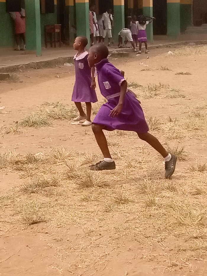 The image captures an outdoor scene at a school in Atani, Nigeria. In the foreground, two children, identified as students by their attire, are visible on a dirt ground scattered with sparse, dry vegetation. The child positioned to the right is in a dynamic, bent-knee posture, indicative of a running or poised-to-run action, facing left. This child wears a purple uniform dress with a white collar, white trim on the cuffs, a white sash, dark closed-toe shoes, and white socks. Directly behind and slightly to her left, a second child stands, also in a purple uniform dress with a white collar, wearing light-colored flat shoes, and looking towards the left.
In the background, a school building with a light-colored facade, green vertical support pillars, and a yellow horizontal stripe along the top of the pillars is visible. Several other children are present near and around the building. One child in a pink garment leans against a green pillar on the far left. Other children in white uniforms are observed standing and one child in white is bent over towards the center-right of the background. A wooden desk or table is discernible inside the building through an opening. The ground is reddish-brown earth, illuminated by natural daylight.