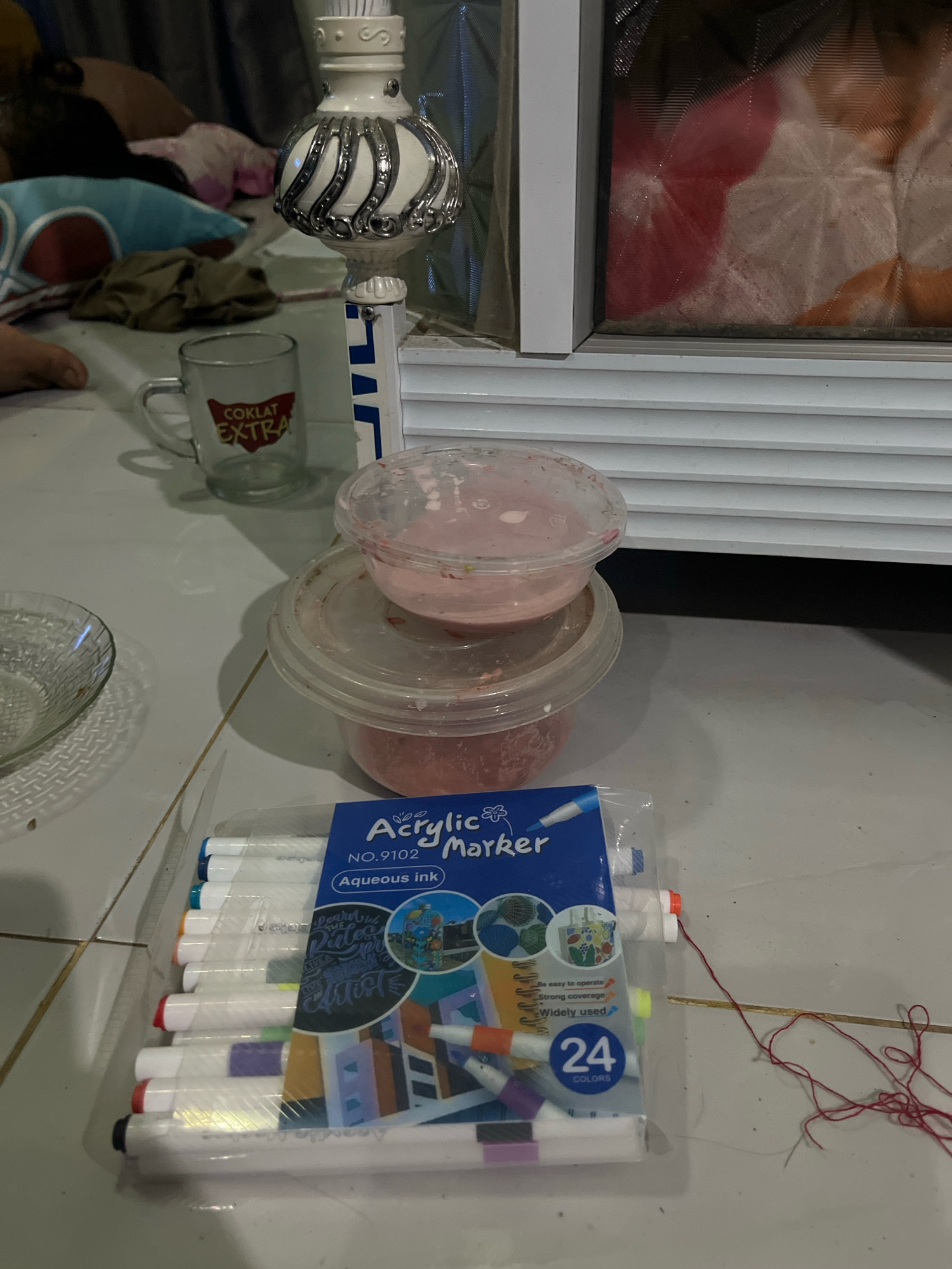 A high-angle, medium shot captures a close-up of a pack of acrylic markers on a tiled surface. The markers are arranged vertically in the foreground, their colorful tips visible through clear plastic packaging. The packaging displays the text "Acrylic Marker," "NO. 9102," "Aqueous ink," and "24 COLORS." To the left of the markers, a clear glass bowl with a woven pattern rests on the tile. Further back, two clear plastic containers, stacked one atop the other, hold a pink substance, possibly food or craft material. A small glass mug with the label "COKLAT EXTRA" is situated behind the containers. In the background, a decorative white and silver object with ornate detailing stands near a white appliance, possibly a microwave. The environment suggests an indoor setting, likely a domestic space in Kebumen, Indonesia. The lighting is dim, casting shadows on the tiled floor.