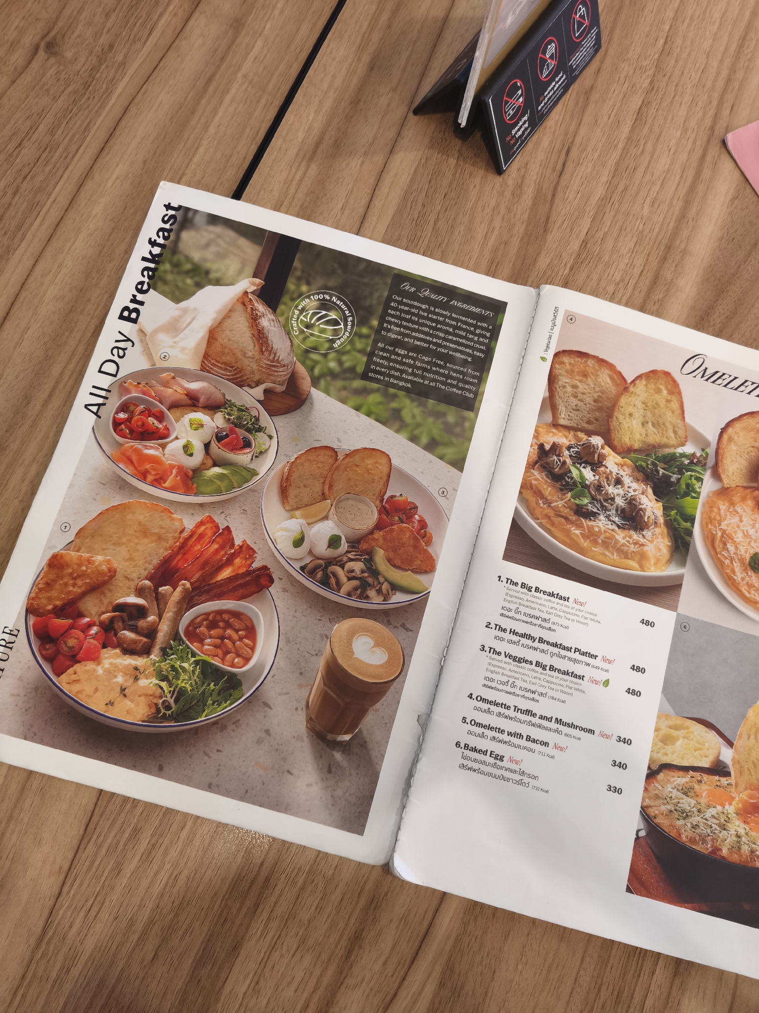 The image displays a menu from a cafe, likely located in Tambon Mai Khao, Thailand. The menu features several breakfast and omelet dishes with accompanying descriptions and prices. In the foreground, there are two plates of breakfast food. The first plate is a full breakfast with eggs, sausages, bacon, baked beans, and vegetables. The second plate contains sliced toast and poached eggs. A latte with latte art is also visible. The background shows images of bread and more food items, and a small information stand with symbols indicating no smoking, no vaping, and free Wi-Fi. The menu itself is placed on a wooden table. The overall aesthetic suggests a contemporary cafe environment.