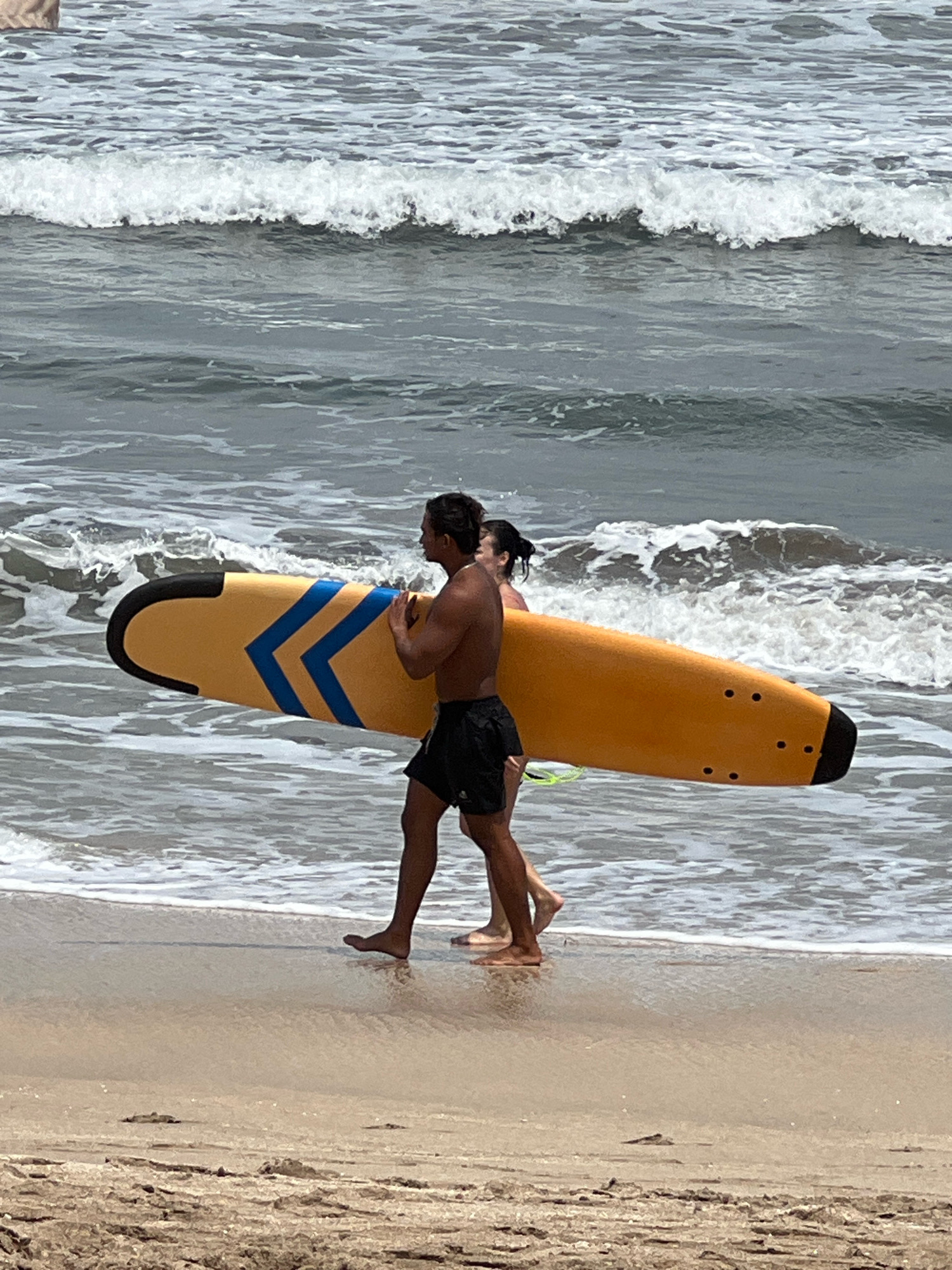 Two individuals, a man and a woman, are walking on a sandy beach in Kuta, Indonesia, near the ocean. The man, shirtless and wearing dark swim trunks, carries a bright yellow surfboard with blue chevron markings and black accents. The woman, visible behind him, appears to be wearing a swimsuit. They are walking towards the right of the frame, with gentle waves lapping at the shore around their feet. The water is a murky gray-blue, reflecting the overcast sky, and white foam from the waves creates a textured line along the beach. The sand is light brown and appears wet near the water's edge, transitioning to drier, coarser sand further up the beach. The background consists of more waves and the open ocean.