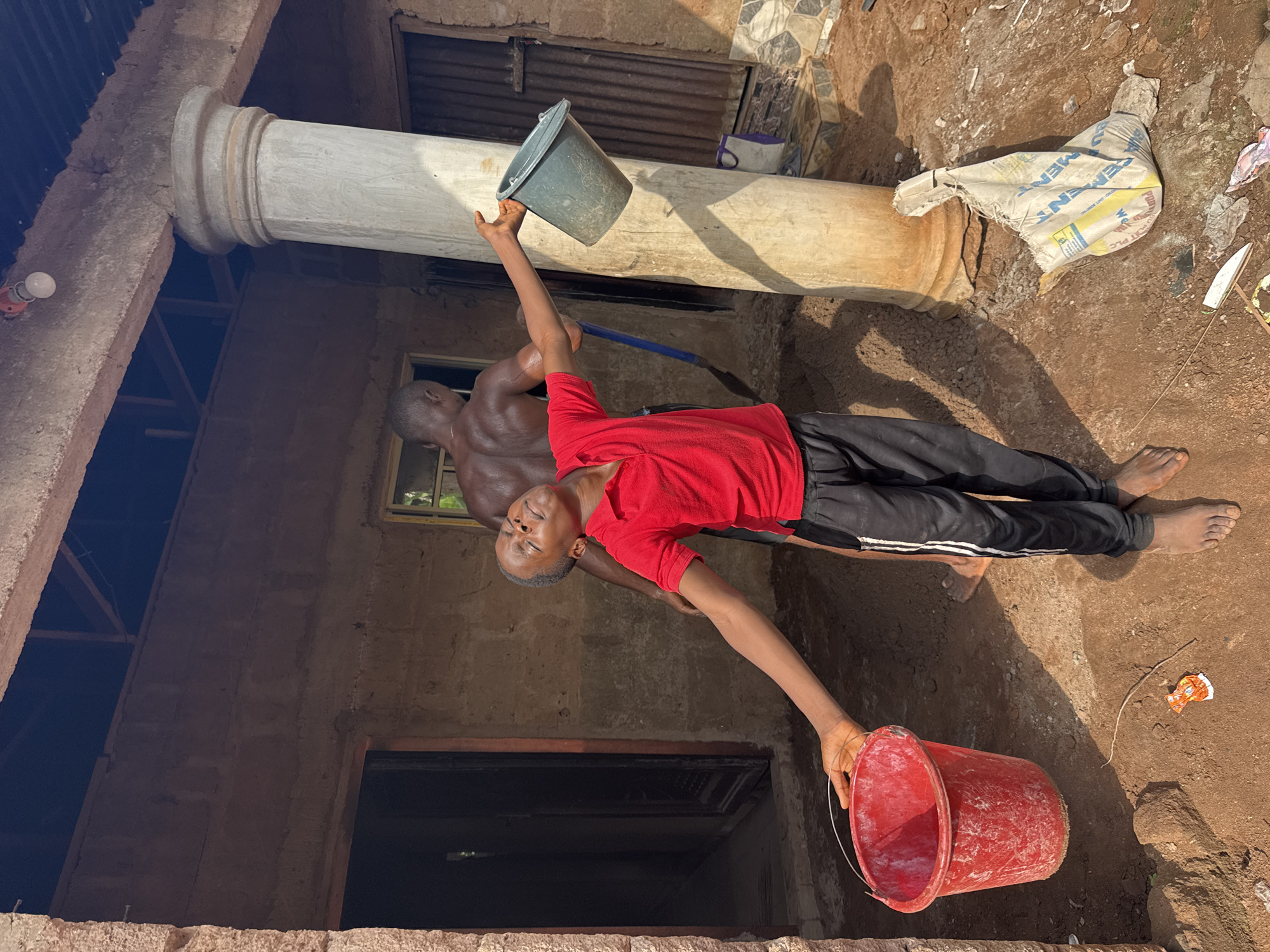In Eziudo, Nigeria, a man in a red t-shirt and black pants holds a red bucket in his outstretched left hand while raising a gray bucket in his right hand. A second man, shirtless, stands behind him. They are near a white, cylindrical object and a ripped cement bag. The ground is dirt, and the surrounding structures appear to be under construction or renovation. A window is visible in the background.