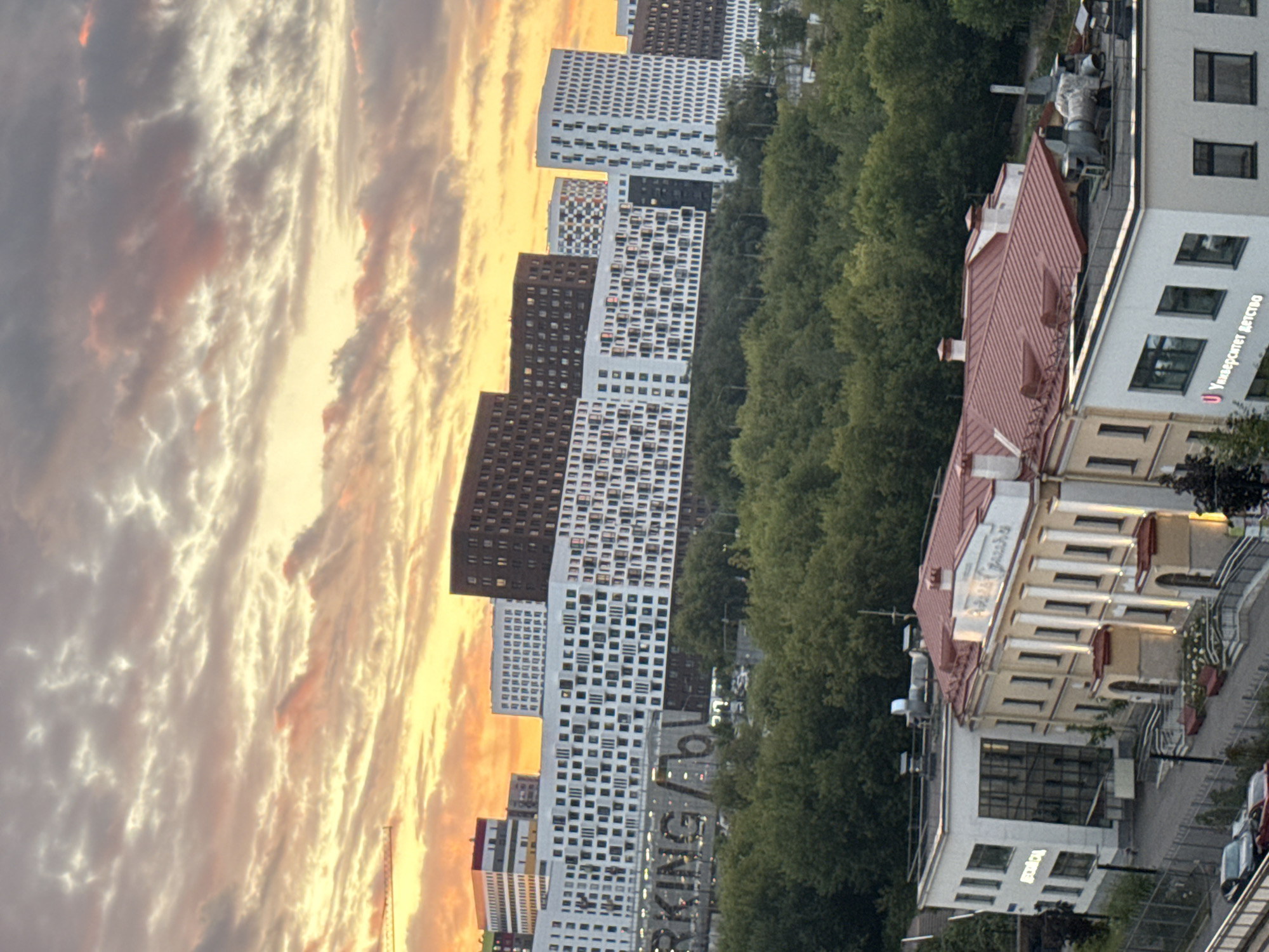 This image depicts a city skyline at sunset, likely in Moscow, Russia, given the architectural style and the Cyrillic signage. The foreground features a cluster of low-rise buildings with red tiled roofs and cream-colored facades. One building has a modern glass entrance, and another displays Cyrillic text that appears to read "Университет Детство" (Children's University). A fenced area with parked cars, including a red car and a dark sedan, is visible in the lower left. The middle ground is dominated by a dense expanse of green trees, suggesting a park or forested area within the city. Behind the trees, a series of modern, multi-story residential buildings stand out. These buildings vary in design, with some featuring predominantly white facades adorned with numerous square windows, while others are darker, possibly brown or grey. The skyline is further punctuated by a few taller, more distinctly shaped buildings. The sky above is filled with dramatic clouds illuminated by the warm hues of a sunset, with oranges and yellows blending with grey and pink tones. The overall scene captures a blend of older and contemporary architecture against a natural backdrop, under a striking twilight sky.