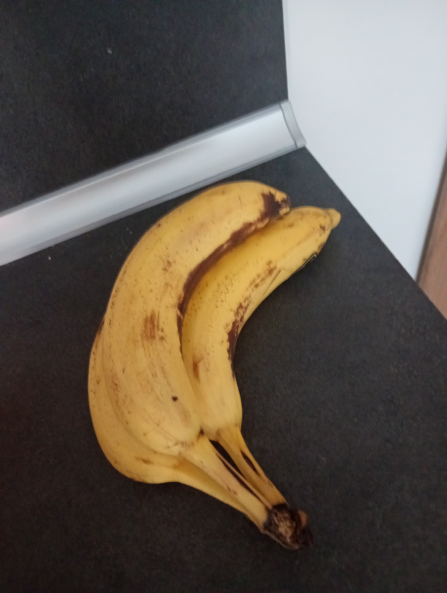 Two bananas are fused together at the stem and lie on a dark, textured surface. The bananas are yellow with brown spots, indicating ripeness. Behind the bananas, a reflective strip runs horizontally across the frame, separating the dark surface from a white vertical surface. The scene is lit from above and to the right. The location is not discernible from the image.