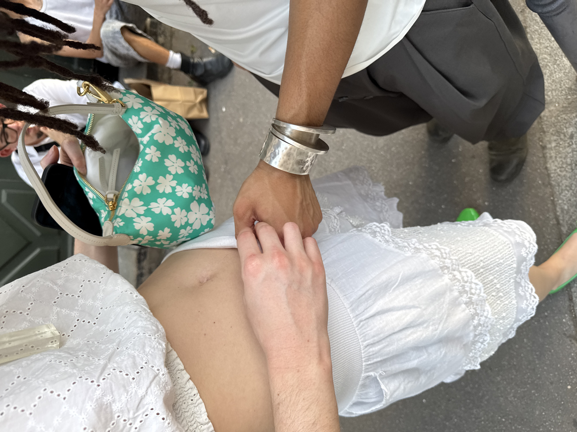 The image captures a scene in what appears to be Paris, France, with the ground showing typical Parisian pavement. Three individuals are partially visible.
One person, likely female, is wearing a white, eyelet-style top and skirt, exposing part of their torso. Another person, positioned above, is wearing a white t-shirt and grey trousers, along with a wide silver bracelet on their right wrist. Their hand is resting on the abdomen of the first person. The third person is wearing glasses, is carrying a small, light green purse with a white floral pattern, and has their hair in dreadlocks.
The overall perspective is from above, looking down at the interaction.