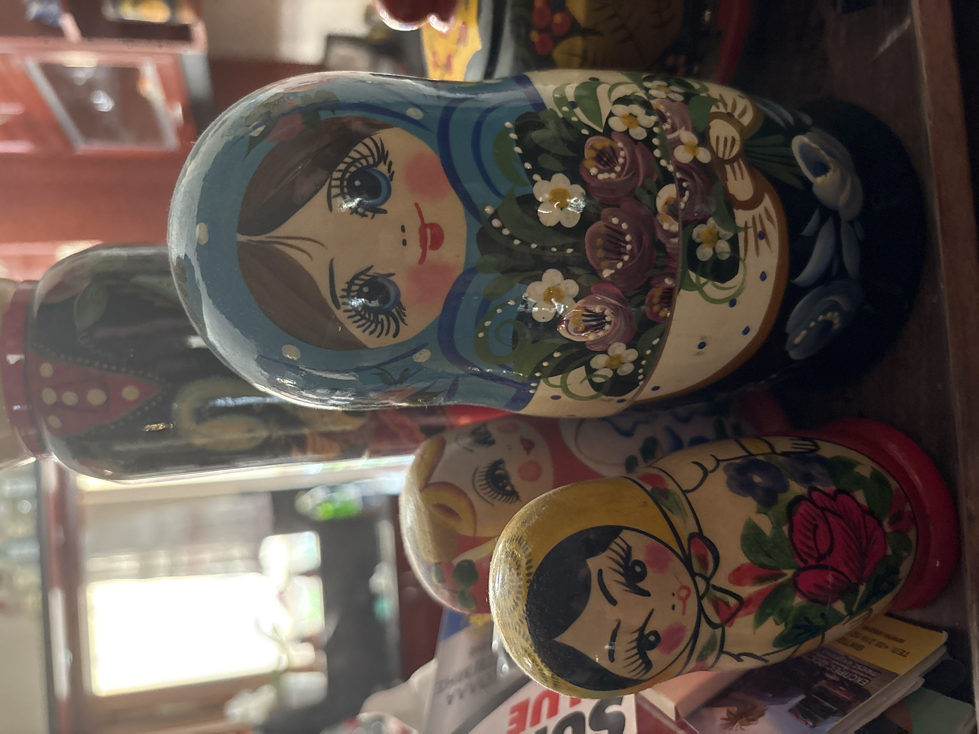 The image shows a collection of Russian nesting dolls, also known as Matryoshka dolls, displayed on a wooden surface. There are at least three dolls visible. The largest doll in the foreground is painted with a blue headscarf, large blue eyes, and is holding a bouquet of flowers, including pink roses and white daisies. To the left and slightly behind it, a smaller doll with blonde hair and a red dress is partially visible. Further to the left and in the foreground, another small doll with a yellow headscarf and a red rose painted on its body is prominently displayed. The background is blurred, but suggests an indoor setting with shelves and objects, possibly in a home or shop. The lighting suggests it is daytime, with light coming from a window. The specific location in Ilion, Greece, is not discernible from the image.
