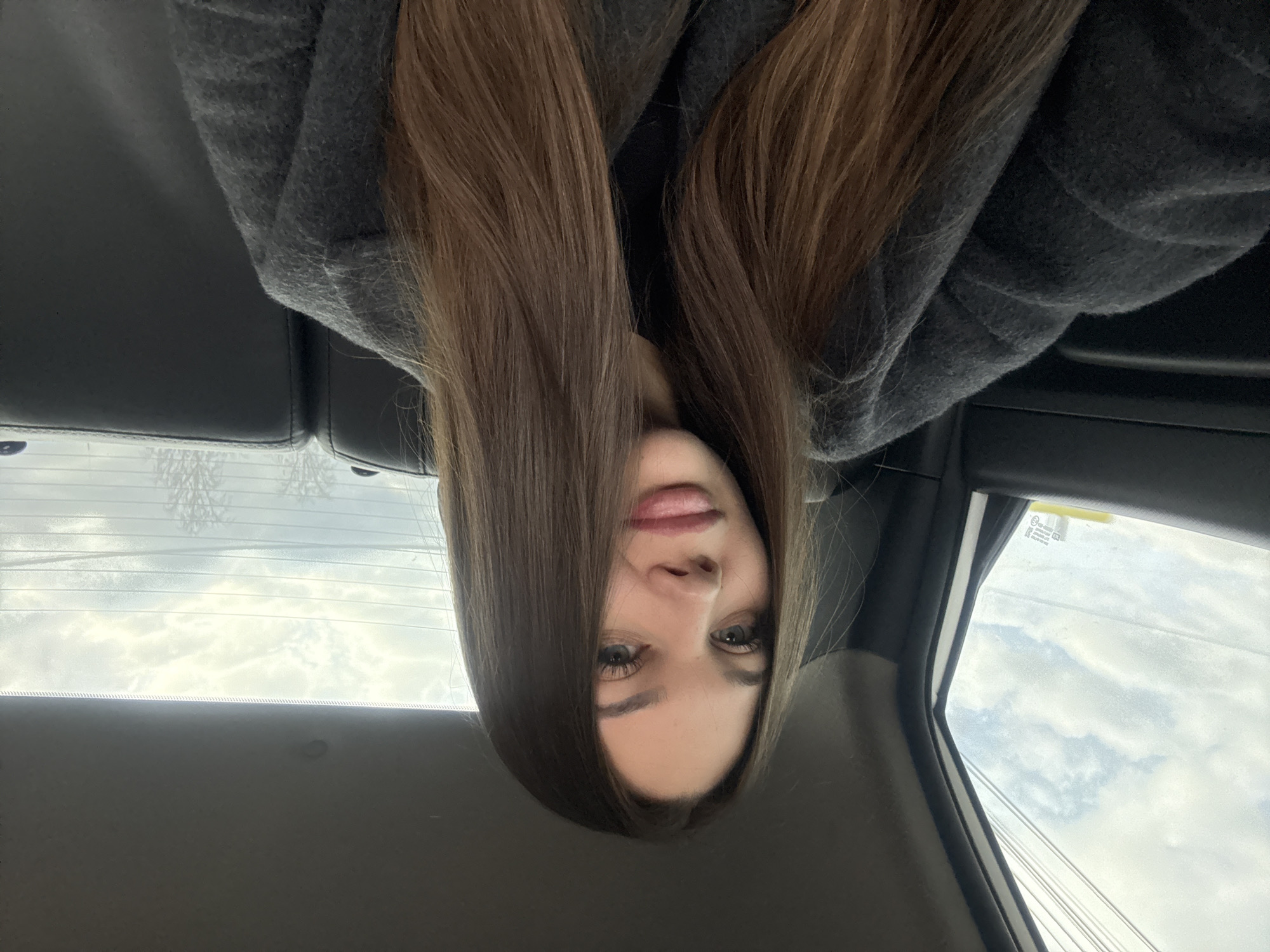 The image depicts a woman inside a car, likely taken from the back seat. She has long, brown hair that falls down over her face and is wearing a gray hooded sweatshirt. The car's interior is visible, including the back seat, rear window with defroster lines, and the side window showing a cloudy sky and power lines. Based on the background details and the prompt's information, the image may have been taken in Chaikovsky, Russia. However, there are no specific identifying features in the image to confirm the exact location.