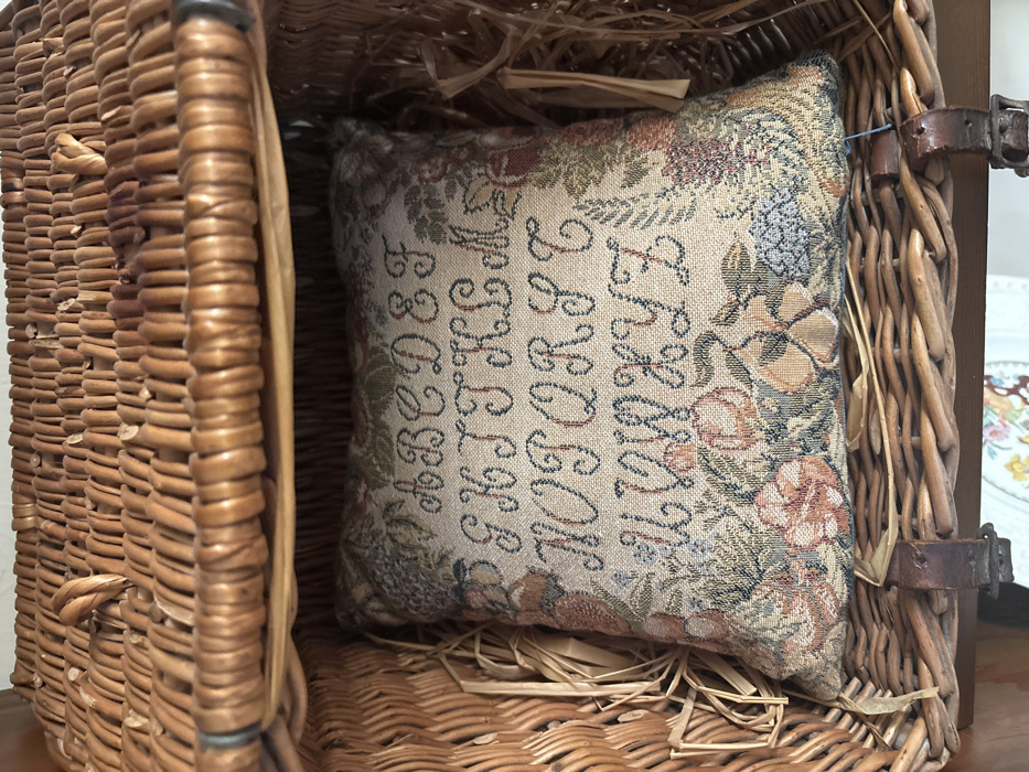 The image displays a woven tapestry-style cushion positioned inside a wicker basket. The cushion, rectangular and plush, features a stitched alphabet from A to Z in uppercase cursive script, arranged in four rows on a light beige background. The letters are dark blue-green. Surrounding the alphabet, and across the bottom of the cushion, is a border of floral and botanical motifs in muted tones of green, rust, light blue, and beige.
The cushion rests within a light brown wicker basket, which appears to be a hamper or trunk given its construction. The interior sides of the basket show additional loose, light-colored straw-like packing material. The upper portion of the image depicts the underside of the basket's lid, also crafted from interwoven wicker. Towards the bottom of the basket, dark brown leather straps with metal buckles are visible, securing the basket's structure or lid. No people are present. The specific location within the city of Monterey, United States of America, is not discernible from the visual information provided in the image.