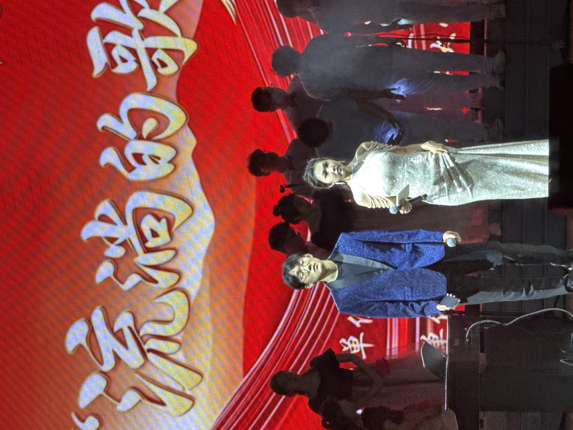 An indoor stage performance is captured in Guta, China, likely during an evening event. The main subjects are two performers on stage, holding microphones. A woman, dressed in a shimmering, light-colored long gown, stands centrally, looking towards the audience. To her right, a man wearing a dark blue patterned jacket, black shirt, and glasses also holds a microphone. Behind them, several other individuals, likely a chorus or backing vocalists, are partially visible in darker clothing.
The backdrop is a large screen displaying vibrant red graphics with prominent, stylized light-colored Chinese characters. This screen serves as a dynamic visual element for the performance. On the right side of the stage, stage equipment such as speakers and stands are visible.
The event is a musical performance or concert, with the central figures actively singing. The large screen and stage setup suggest a professional production. Visible text on the backdrop reads "奋进的色彩" (Fènjìn de sècǎi), which translates to "Colors of Progress" or "Vibrant Colors of Striving." Below this, the text "2023 年" (2023 Nián) is also discernible, indicating the year 2023.