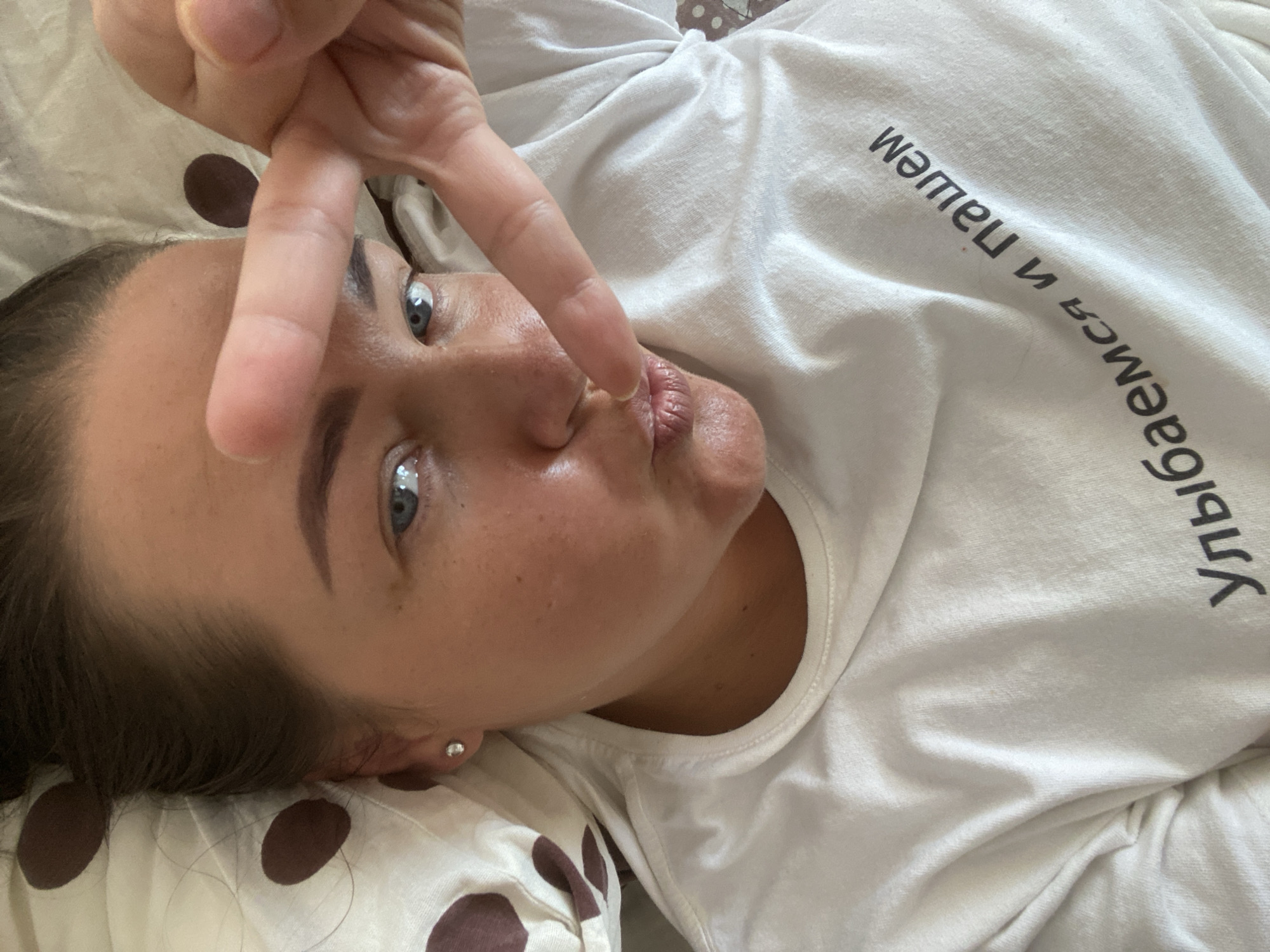 A close-up, overhead selfie shows a person lying down, wearing a white t-shirt with Russian text printed on it. The text reads "Улыбаемся и Пашем", which translates to "We smile and work". The person is making a peace sign with their right hand, covering part of their forehead. They have dark hair pulled back and blue eyes. The background appears to be a pillow with a brown polka dot pattern. There are no discernible landmarks or elements that would indicate a specific location within the city of Sochi, Russia. The image is well-lit, suggesting it was taken indoors during the daytime.
