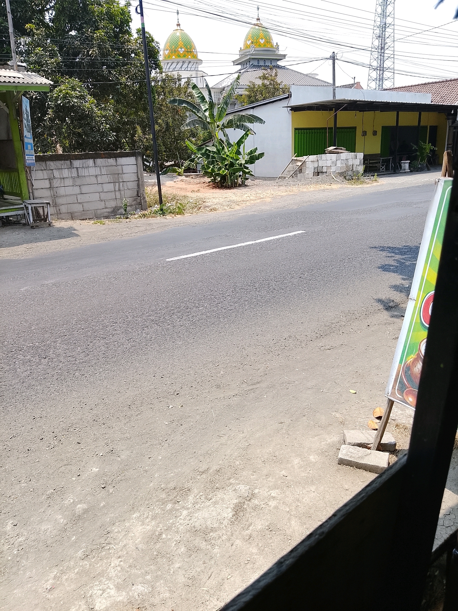 The image depicts a street scene in Banaran, Indonesia, on a clear, sunny day. In the background, a mosque with green and yellow domes is visible, along with a modern building with a yellow facade and a black awning. A transmission tower stands tall in the distance. In the foreground, a paved road runs through the scene, with a white line marking the center. To the left of the road, there's a low concrete wall and some greenery, including banana trees. A roadside stall or shop is partially visible on the left. On the right side of the image, a sign with a green border and a colorful illustration is propped up by bricks. The ground is a mixture of dirt and gravel. There are no people visible in the frame.