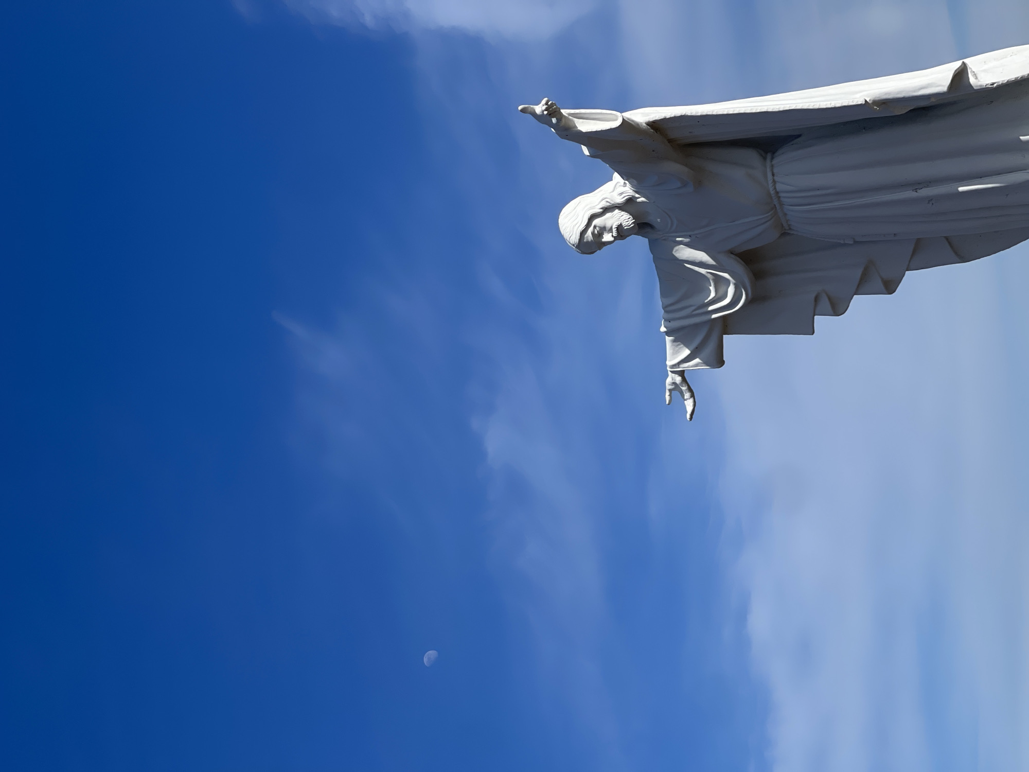 The image depicts the statue of Christ the King in Krasnoyarsk, Russia, against a backdrop of a blue sky with some wispy clouds. The statue is white and shows Christ with his arms outstretched. A crescent moon is visible in the lower left portion of the image. The photograph has been rotated 90 degrees counterclockwise.