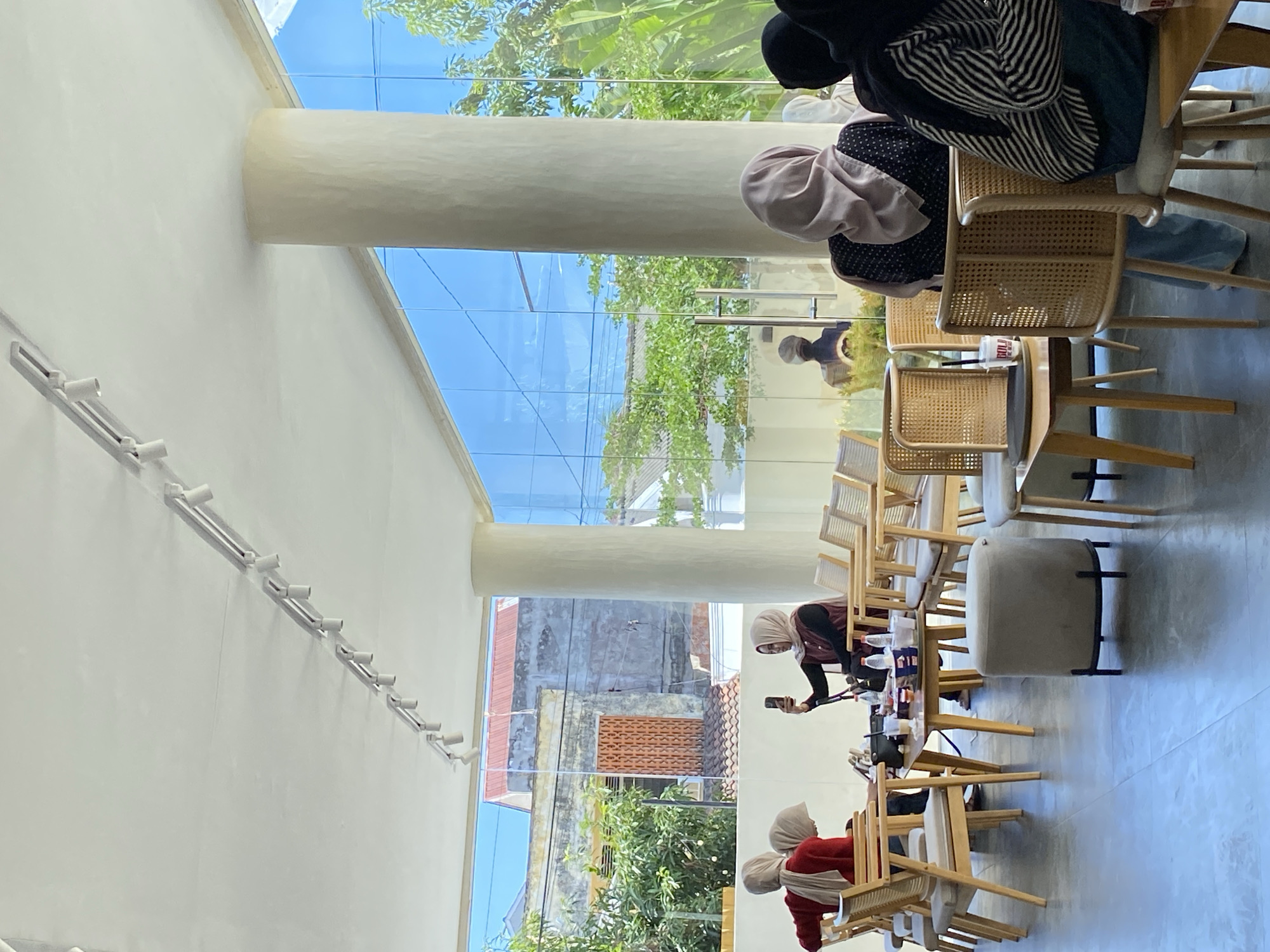 The image depicts the interior of a cafe in Lhokseumawe, Indonesia. The space features a glass ceiling and large pillars that allow natural light to filter in. The seating arrangement includes wooden tables and chairs, occupied by individuals wearing hijabs. One person is seen holding a smartphone. The outside view through the glass ceiling and windows includes buildings and greenery.