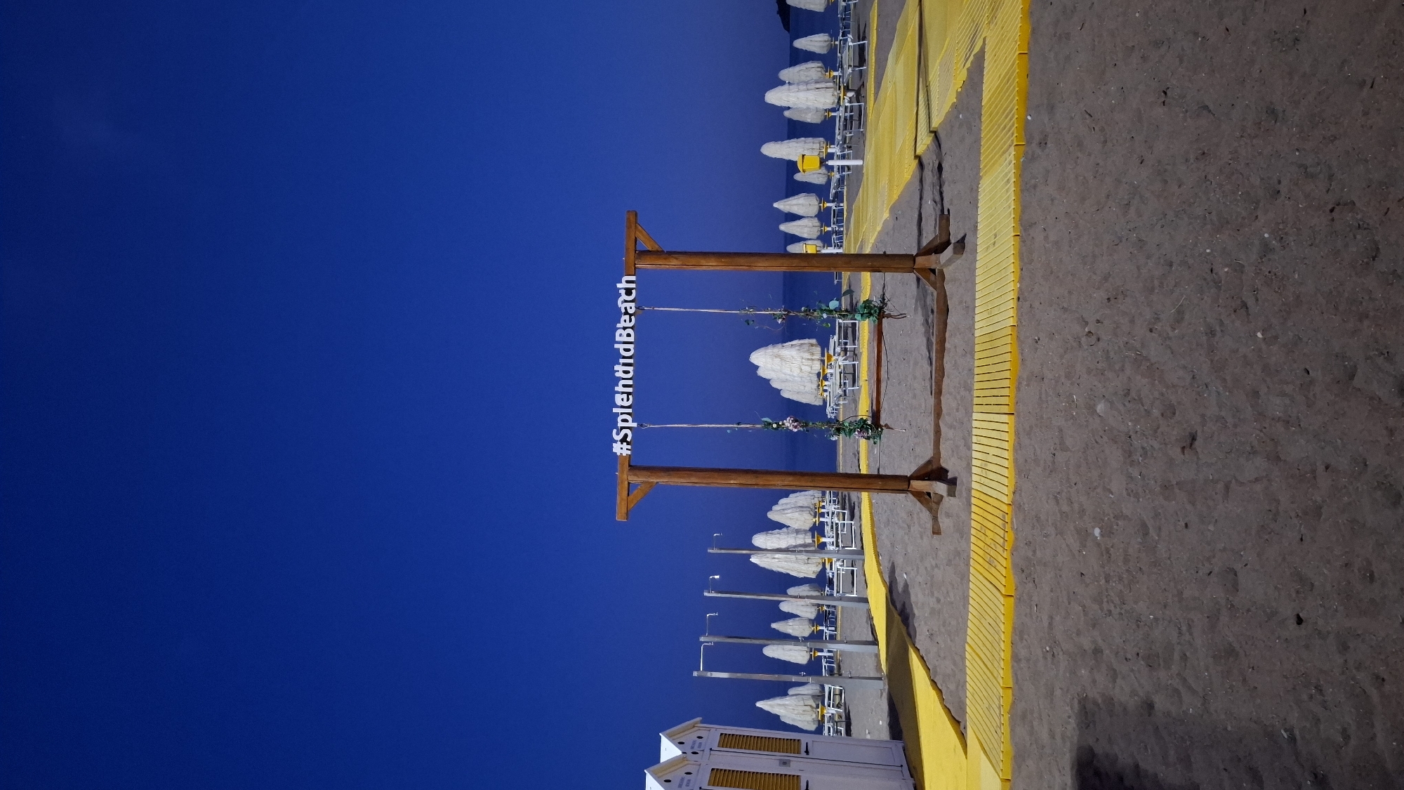 This image captures a tranquil beach scene at what appears to be dusk or night in Boreti, Montenegro. The sky is a clear, deep blue, indicating the late hour. In the foreground, there is a sandy beach. A notable wooden structure, resembling a swing frame or a photo prop, stands on the sand. White letters on its horizontal beam spell out "#SplendidBeach". Greenery and delicate flowers are used as decorations, hanging from the ropes of the swing.
Parallel to the shoreline, a distinct yellow wooden walkway or path stretches along the beach. Adjacent to this pathway, rows of neatly arranged white beach umbrellas and sun loungers are visible, all facing towards the sea. In the lower left corner of the image, a white beach hut with yellow slatted windows and doors is partially seen. The calm water of the sea extends into the distance, meeting the horizon under the vast blue sky. The scene evokes a peaceful and well-maintained resort beach atmosphere.
