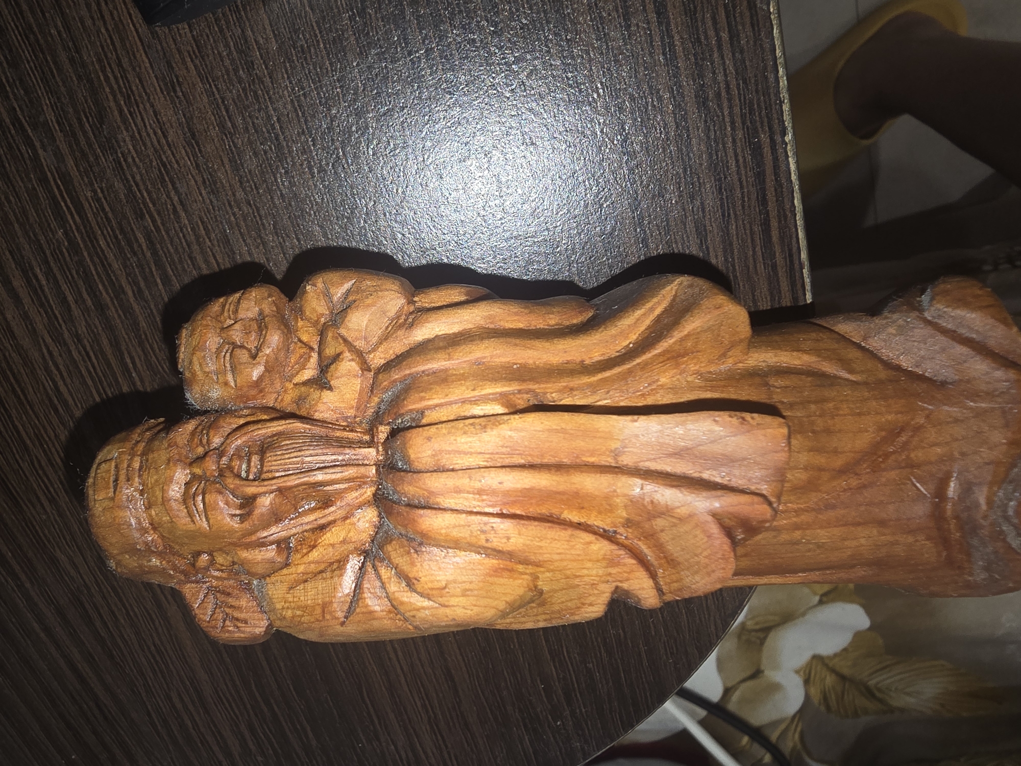 The image is a close-up, top-down view of a carved wooden figurine of an elderly man holding a child. The figurine is made of light-brown wood with visible grain and some darker accents. The elderly man has a long, flowing beard and is smiling with his eyes closed. The child, also smiling, is nestled in the man's arms. Both figures are carved with simple, rounded features. The figurine is positioned on a dark brown, wooden surface with a visible wood grain pattern. The lighting creates a shadow to the left of the figurine. The background is out of focus, showing a floral pattern on fabric to the left and a dark, textured surface with a bright reflection on the right. There is no information in the image to suggest a specific location in Ulan-Ude, Russia.
